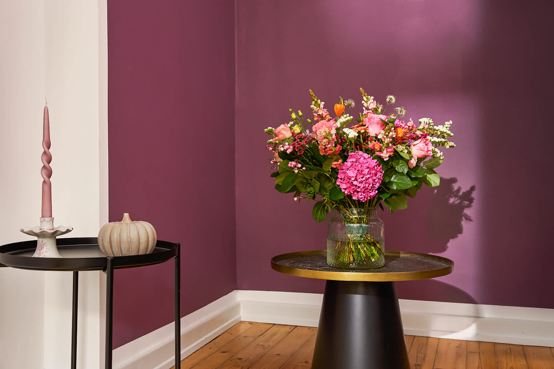 Ein farbenfroher Strauß rosa und gelber Blumen steht auf einem Holztisch vor einer dunkelvioletten Wand, im Hintergrund sind eine Kerze und andere dekorative Elemente zu sehen.
