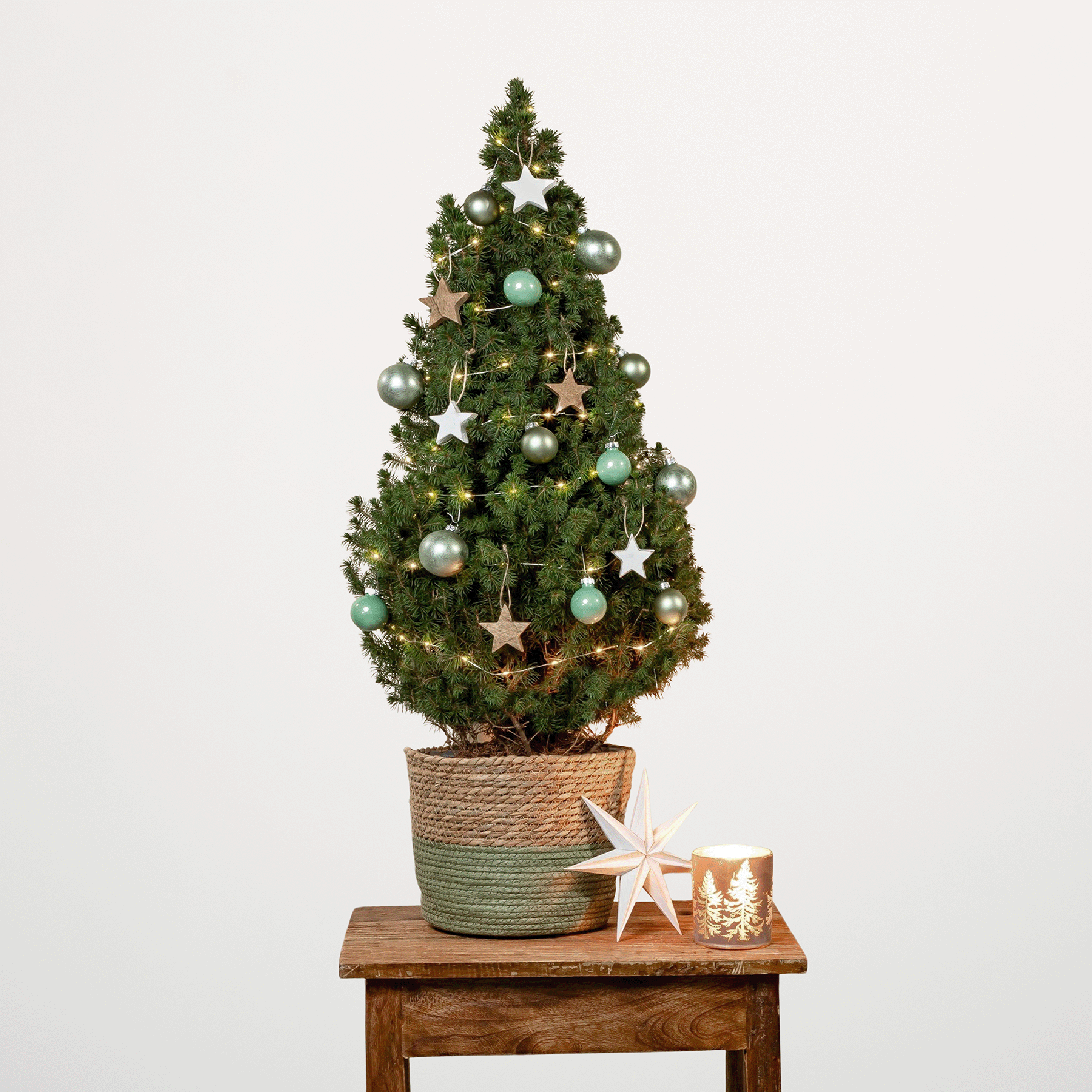 Auf einem Holztisch steht ein kleiner, geschmückter Weihnachtsbaum, umgeben von festlichem Schmuck und einem sternförmigen Licht, vor einem schlichten beigen Hintergrund.
