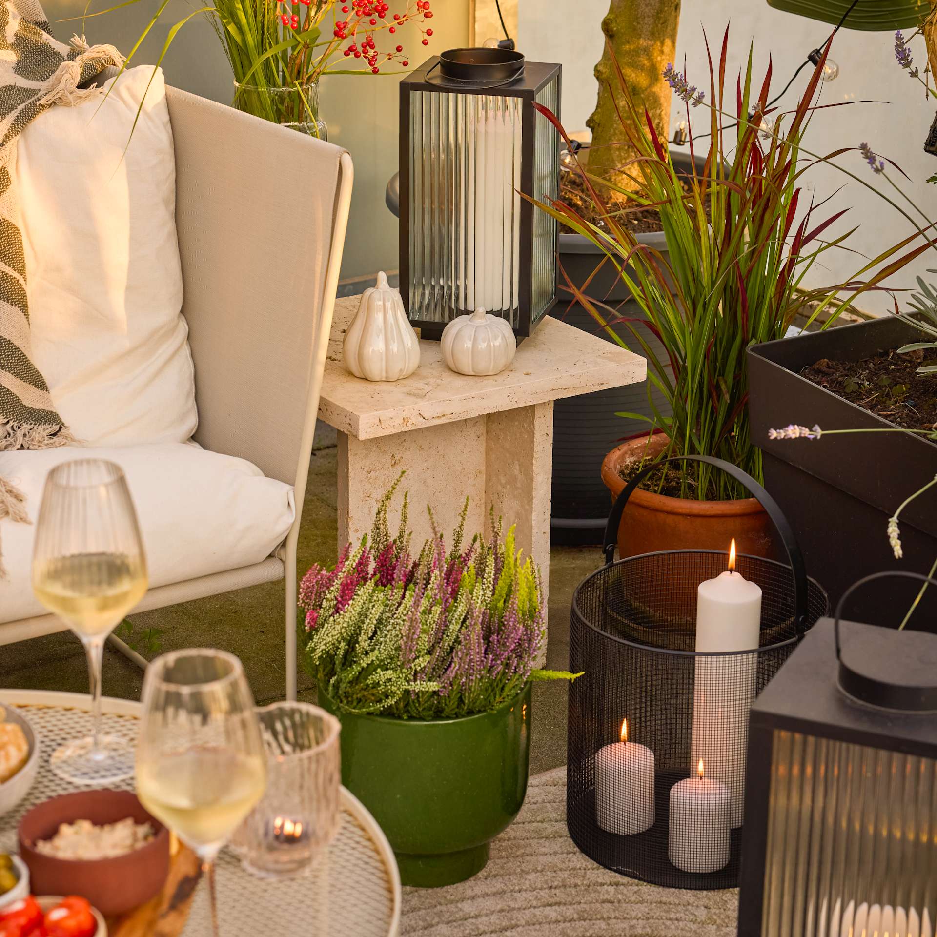 Ein gemütlicher Sitzbereich im Freien mit verschiedenen Dekorationselementen, darunter Topfpflanzen, Kerzen und ein Glas Wein auf einem Holztisch.