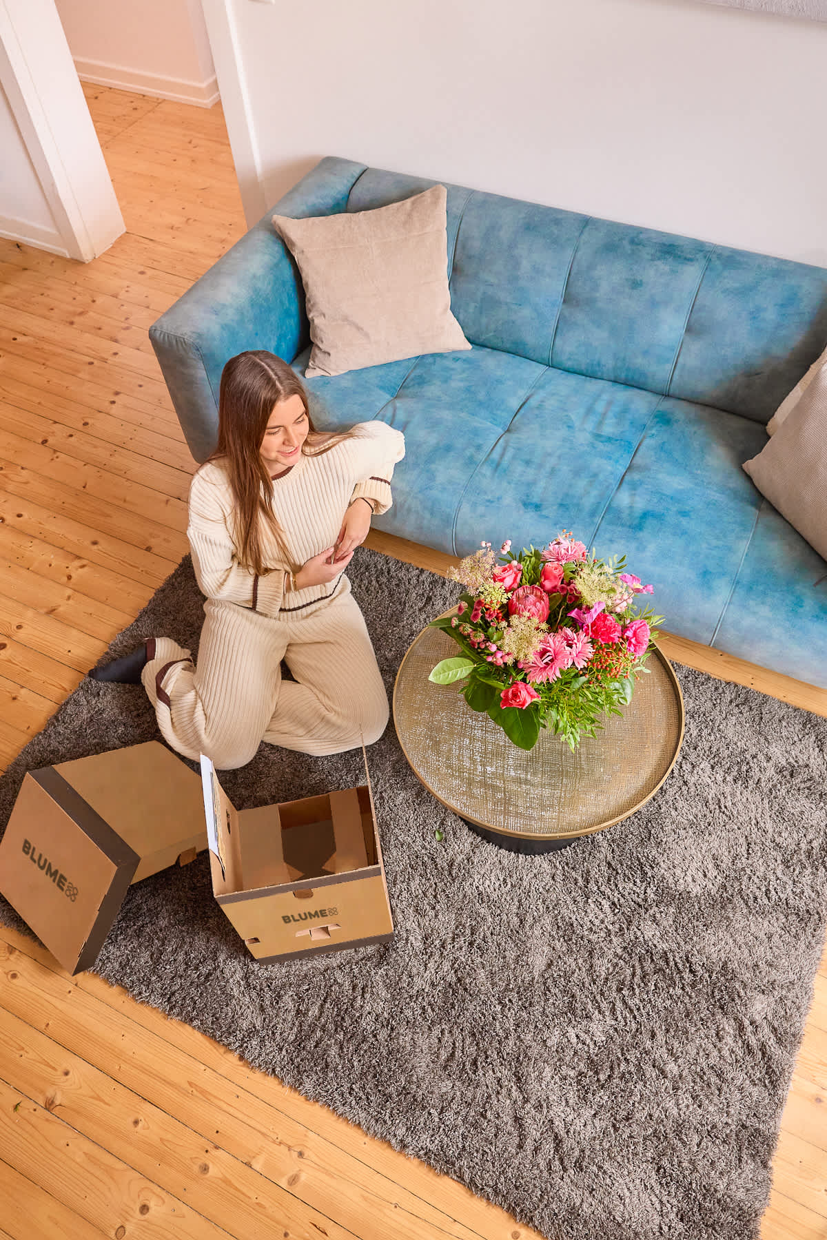 Eine Frau in einem kuscheligen Pullover sitzt auf einem plüschigen blauen Sofa, umgeben von einem hölzernen Couchtisch mit einem Blumenstrauß und Pappkartons, in einem warmen und einladenden Wohnzimmerambiente.
