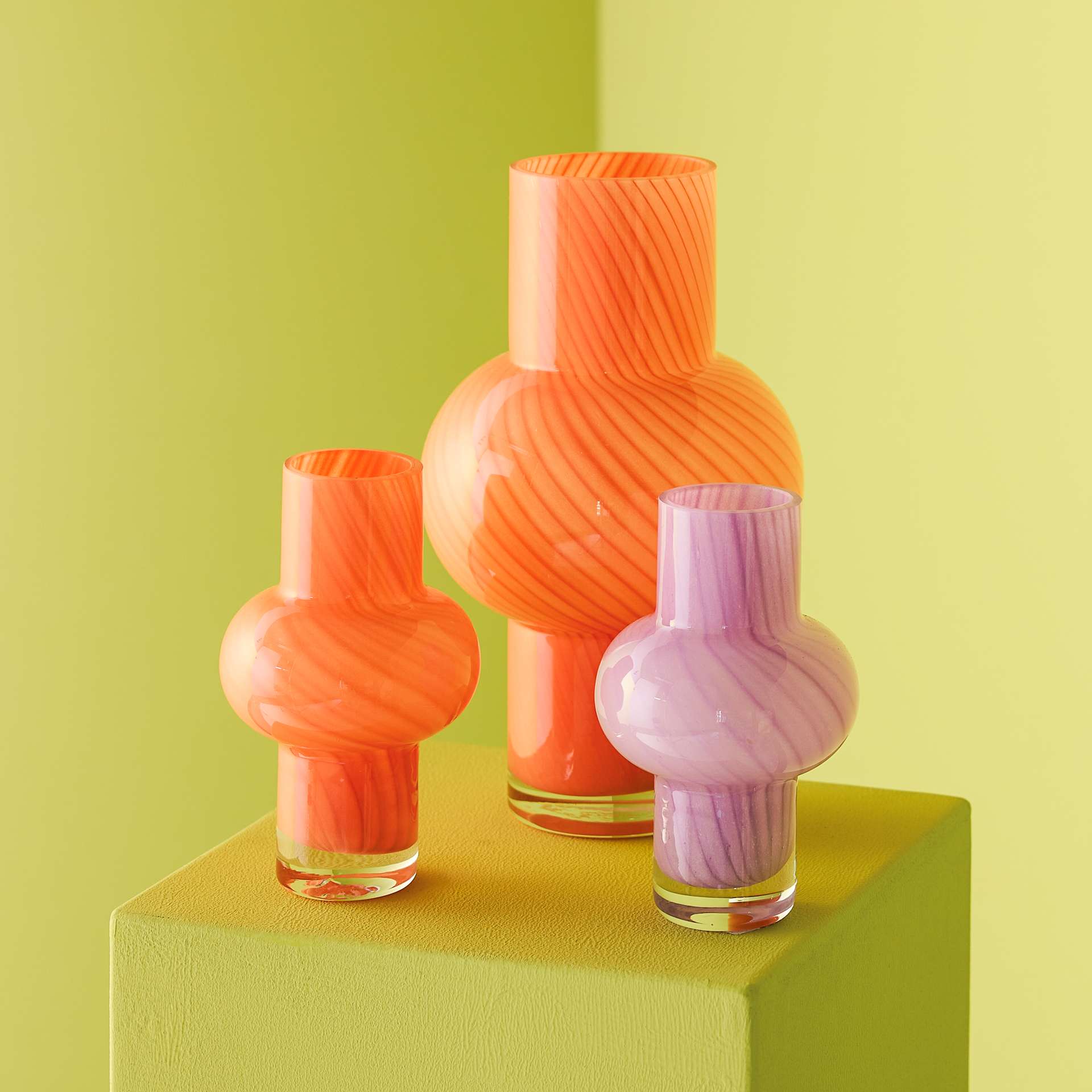 Drei leuchtende Glasvasen in verschiedenen Orange- und Rosatönen stehen auf einem grünen Würfel vor einem leuchtend gelben Hintergrund.