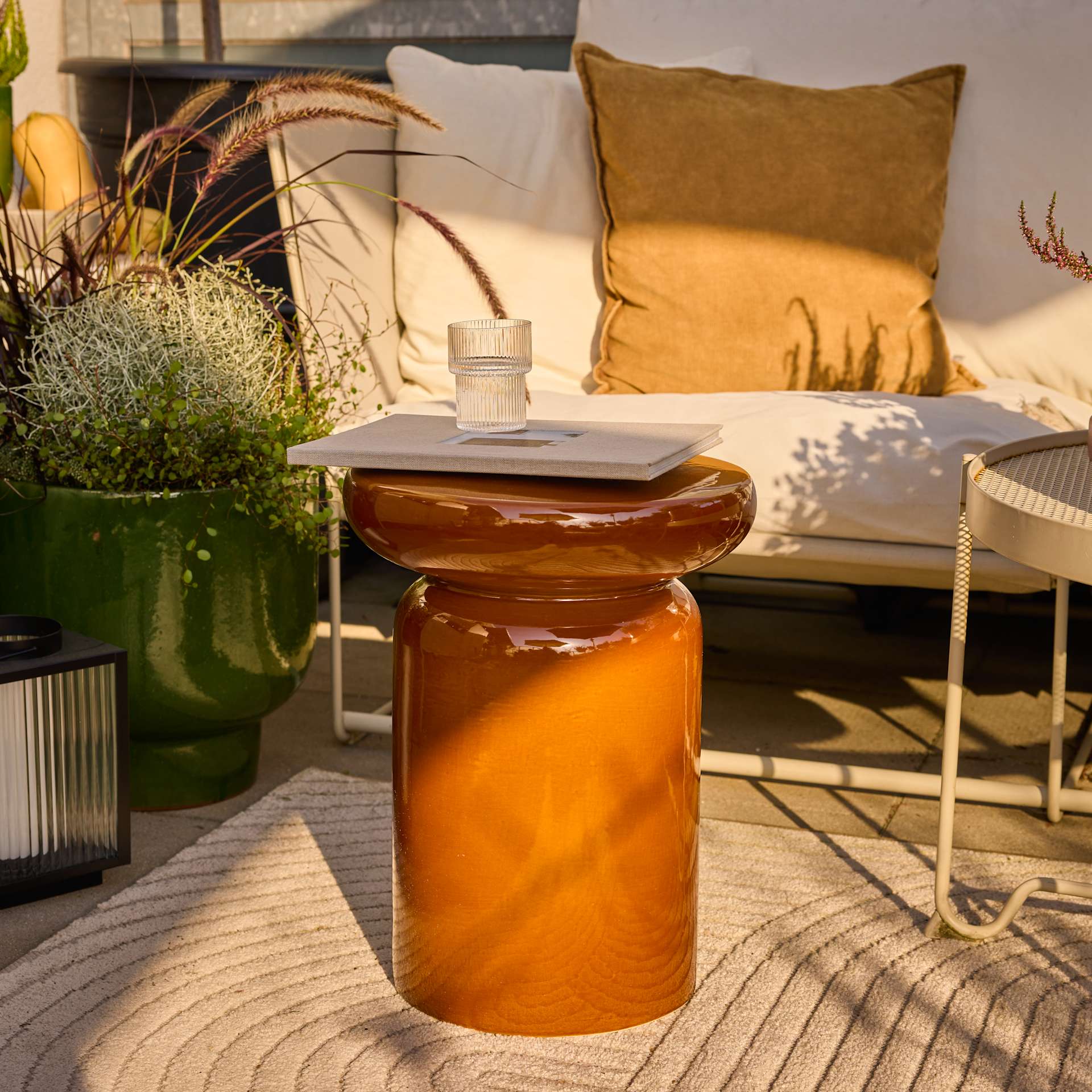 Ein gemütlicher Sitzbereich im Freien mit einem Glasgefäß, das mit einer bernsteinfarbenen Flüssigkeit gefüllt ist, umgeben von dekorativen Kissen, Pflanzen und anderen Akzenten, die eine warme und einladende Atmosphäre schaffen.