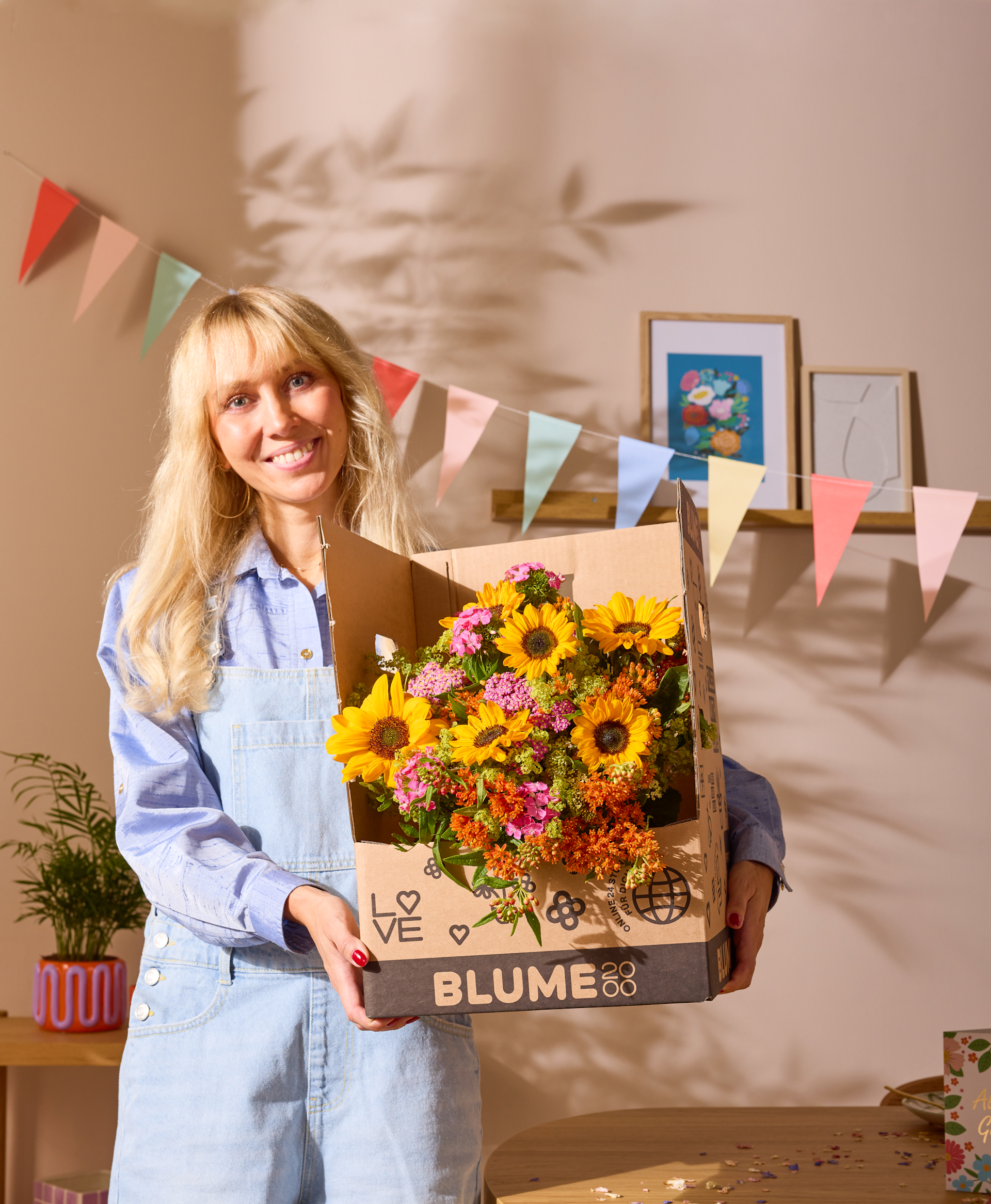 Eine lächelnde junge Frau mit einer Schachtel bunter Blumen steht in einem mit festlichen Fahnen und Kunstwerken geschmückten Raum.