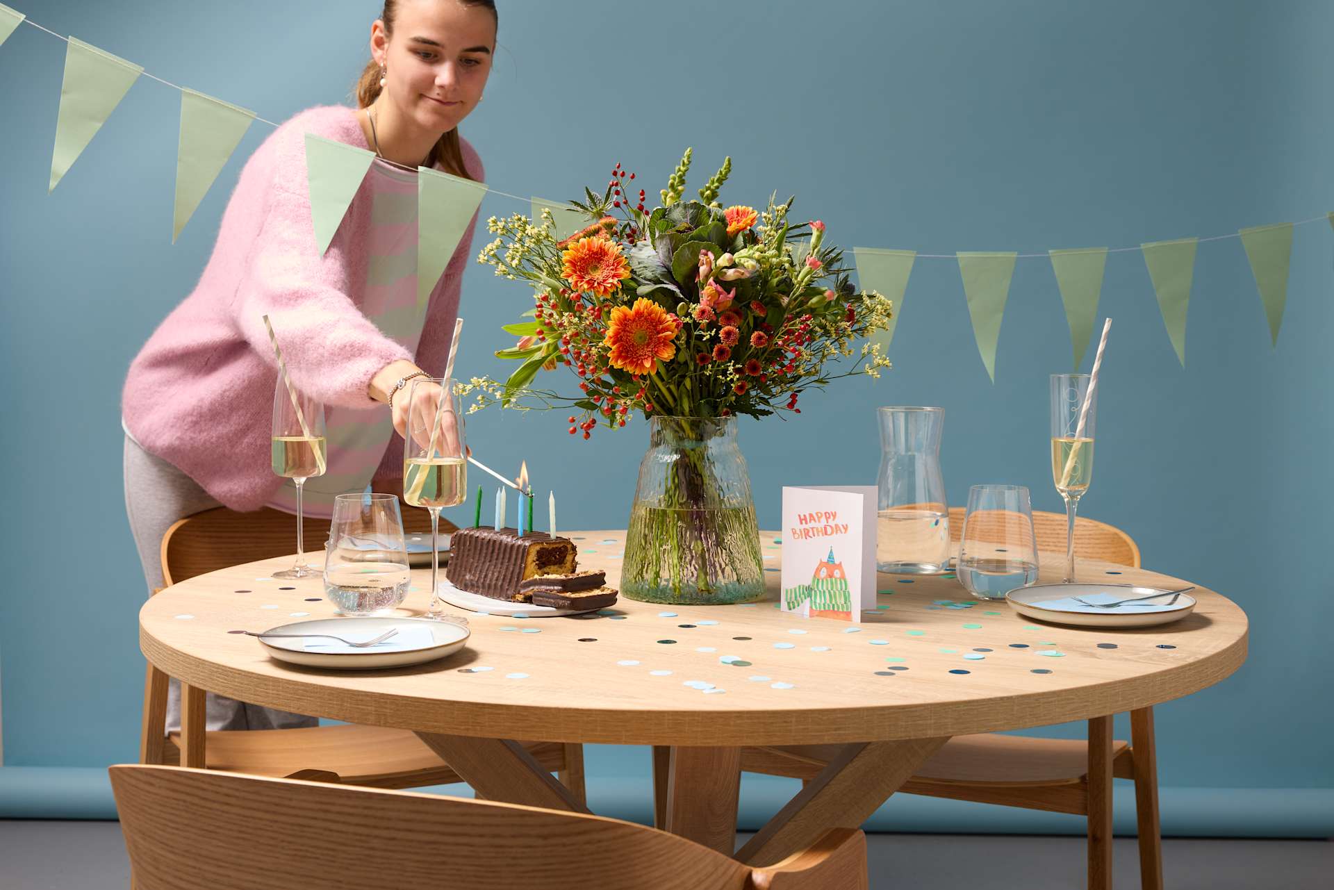 Ein festlich gedeckter Tisch mit einem Strauß leuchtender Blumen, Champagnergläsern und einer Person in einem rosa Pullover, die im Hintergrund vor einer pastellfarbenen, mit bunten Fahnen geschmückten Wand steht.