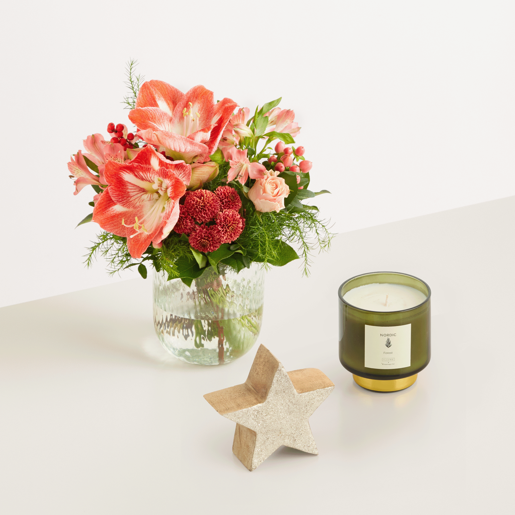 Auf einem Tisch steht ein farbenfroher Strauß roter und rosa Blumen in einer Glasvase, begleitet von einer Kerze und einer sternförmigen Holzdekoration.