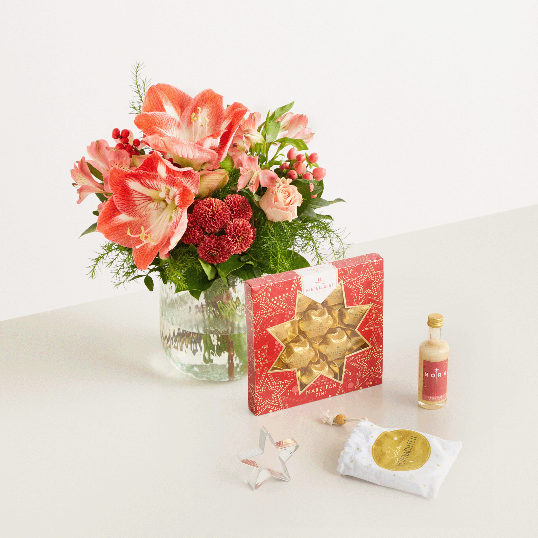 Ein farbenfroher Strauß roter und rosa Blumen, darunter Lilien und Dahlien, steht in einer Glasvase neben einer roten Geschenkbox mit goldener Schleife, einer kleinen Glasflasche und einem weißen Tuch auf einem schlichten Hintergrund.