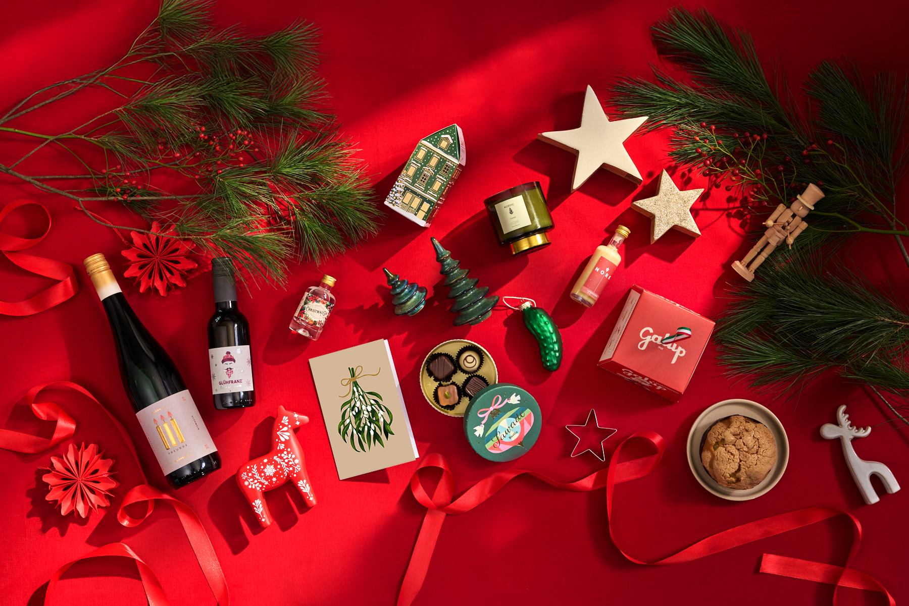 Ein festliches Arrangement weihnachtlicher Gegenstände, darunter Weinflaschen, Ornamente und andere dekorative Elemente, vor einem leuchtend roten Hintergrund.