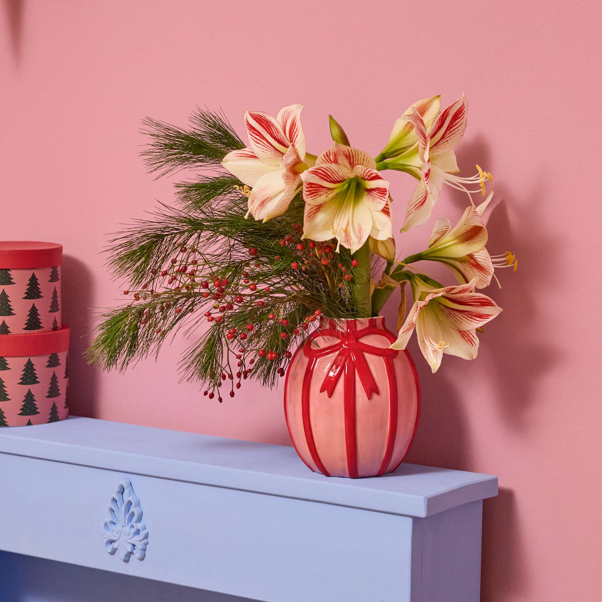 Ein leuchtender Strauß rosa und gelber Blumen steht in einer roten Vase auf einem blauen Tisch vor einem rosa Hintergrund, im Hintergrund ist ein gemusterter Behälter zu sehen.