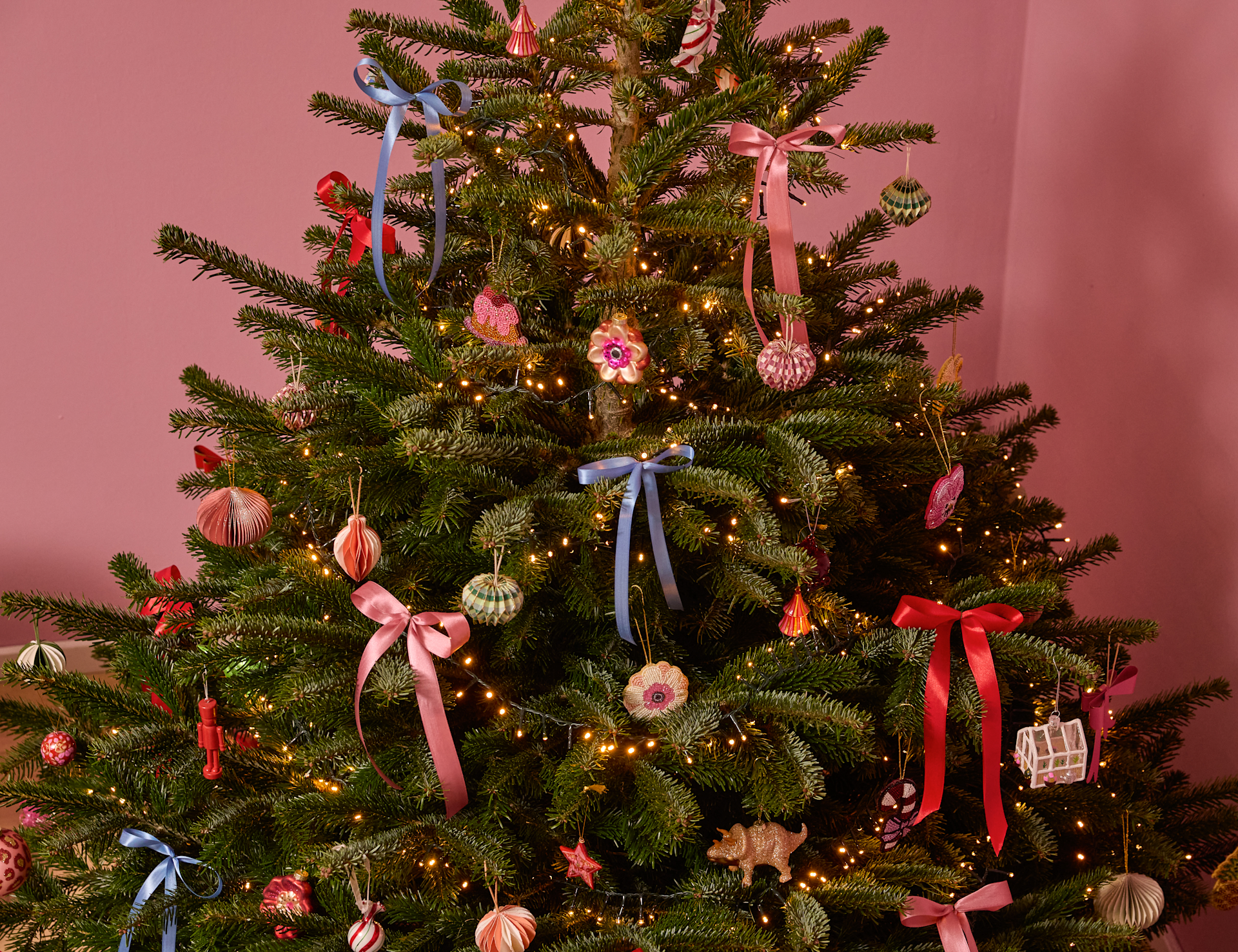 Im Vordergrund steht ein wunderschön geschmückter Weihnachtsbaum, geschmückt mit bunten Ornamenten, Bändern und funkelnden Lichtern vor einem rosa Hintergrund.