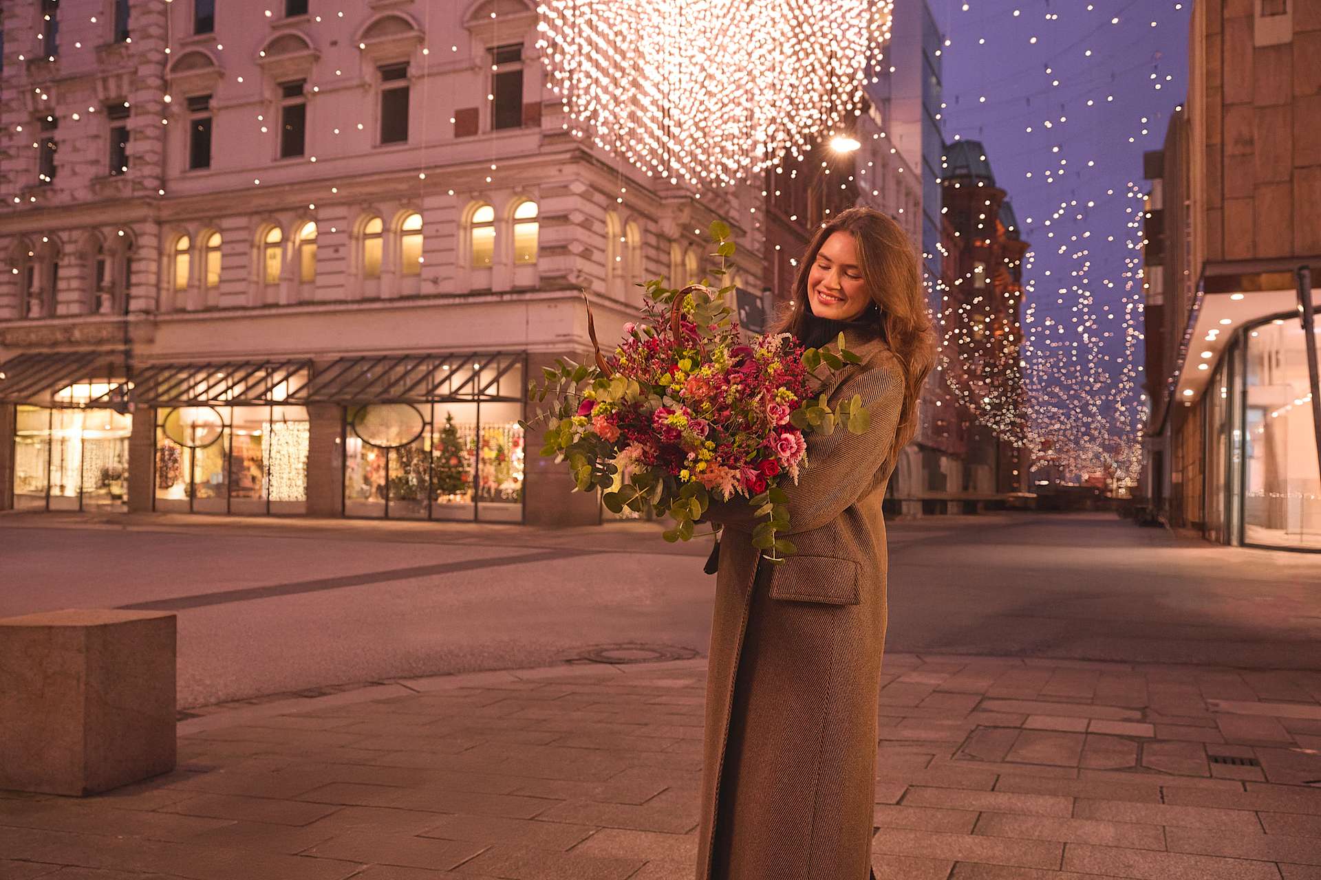 Eine Frau in einem beigen Mantel steht nachts mit einem Blumenstrauß in der Hand auf einer Stadtstraße, während die Gebäude im Hintergrund mit festlichen Lichtern und Dekorationen geschmückt sind.