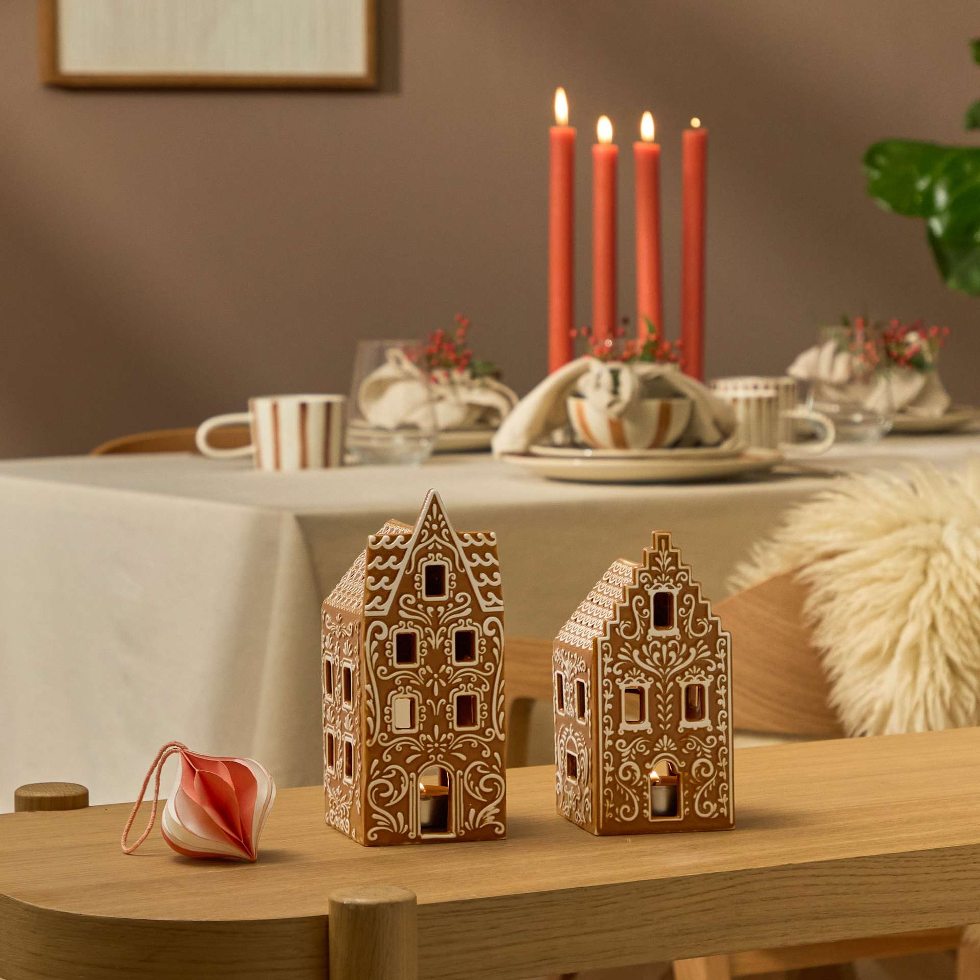 Das Bild zeigt eine gemütliche und festlich gedeckte Tafel mit einem Holztisch, der mit dekorativen Holzhäuschen, roten Kerzen und anderen weihnachtlichen Accessoires geschmückt ist. Im Hintergrund sind ein gerahmtes Kunstwerk und eine Topfpflanze zu sehen, die eine warme und einladende Atmosphäre schaffen.