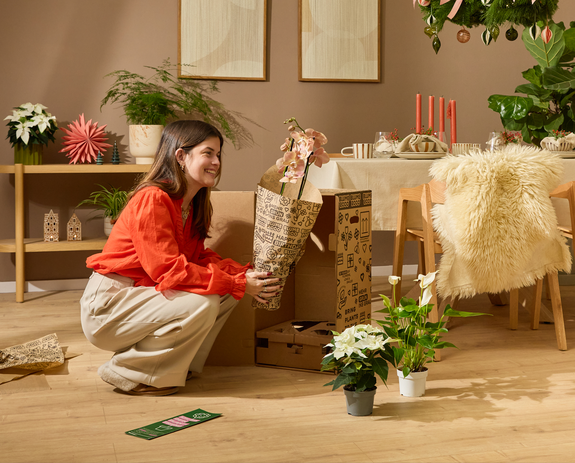 Eine junge Frau in einer roten Bluse sitzt auf dem Boden, umgeben von verschiedenen Dekorationsgegenständen wie Pflanzen, Kerzen und einem flauschigen Sessel, in einer gemütlichen und festlichen Atmosphäre in Innenräumen.
