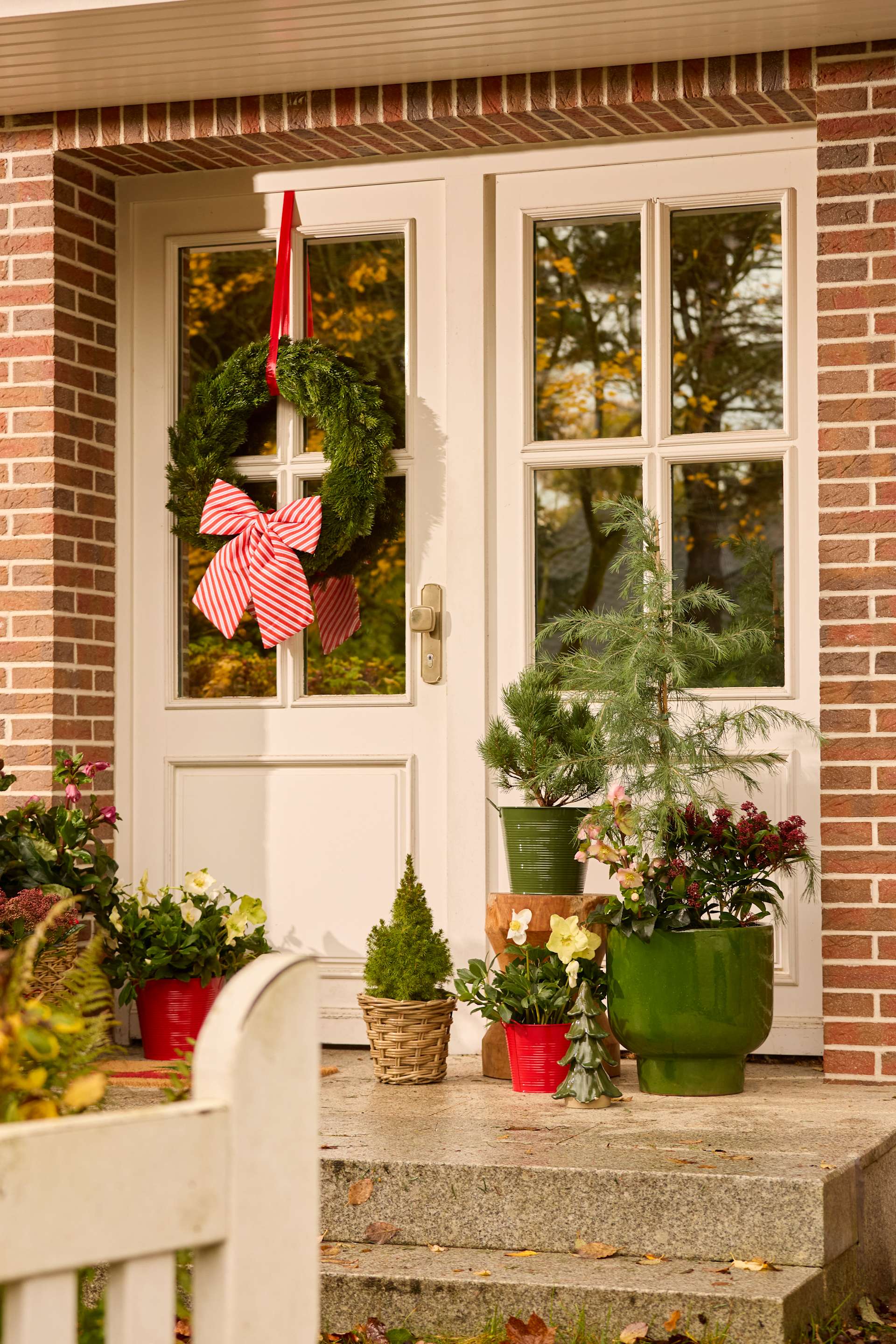 Eine gemütliche und festliche Veranda mit einem Kranz, Topfpflanzen und einer Holzbank vor dem Hintergrund eines Backsteingebäudes und herbstlichem Laub.