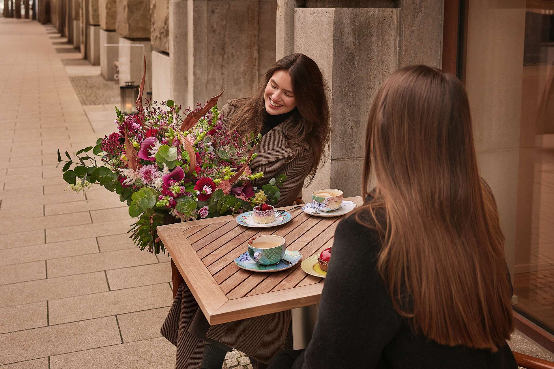 Zwei junge Frauen mit langem, dunklem Haar sitzen an einem Holztisch und genießen eine Tasse Kaffee oder Tee. Der Tisch ist mit einem farbenfrohen Blumenstrauß geschmückt, der dem Bild einen Farbtupfer verleiht. Im Hintergrund bilden Holzsäulen eine Reihe von Säulen, die eine gemütliche und einladende Atmosphäre schaffen.
