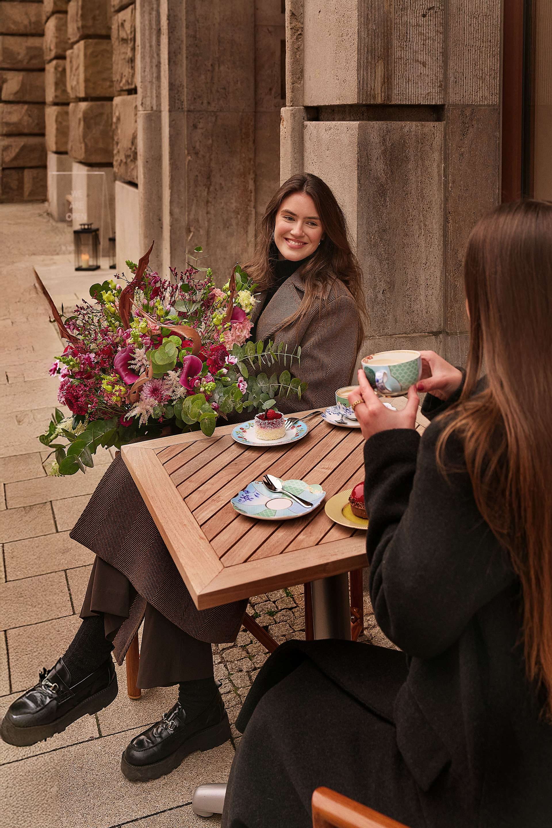 Zwei Frauen sitzen an einem Tisch im Freien und genießen eine Mahlzeit und angeregte Unterhaltung. Der Tisch ist mit einem farbenfrohen Blumenstrauß geschmückt, der dem Bild einen Farbtupfer verleiht. Im Hintergrund ist ein Steingebäude zu sehen, was auf ein urbanes Umfeld hindeutet.