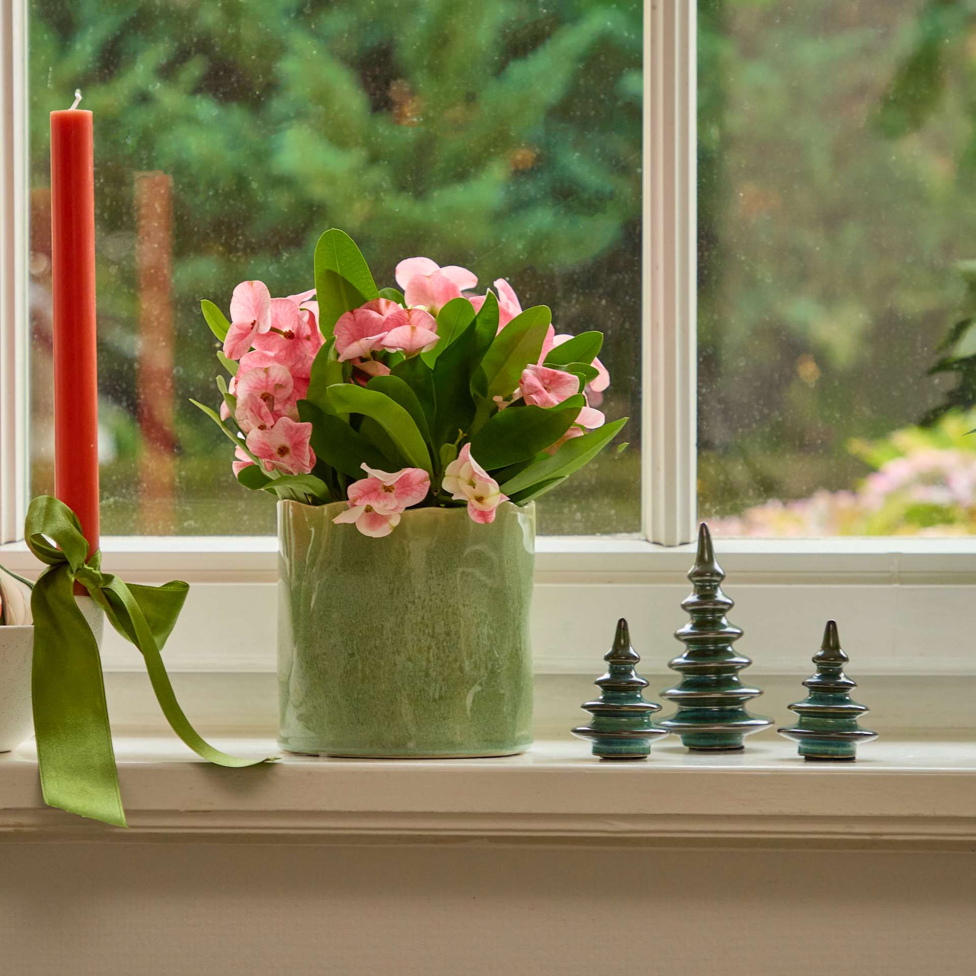 Auf einem Fensterbrett steht eine Vase mit rosa Blumen, im Hintergrund sind eine rote Kerze und dekorative Keramikbäume zu sehen, die den Blick auf eine üppig grüne Landschaft freigeben.