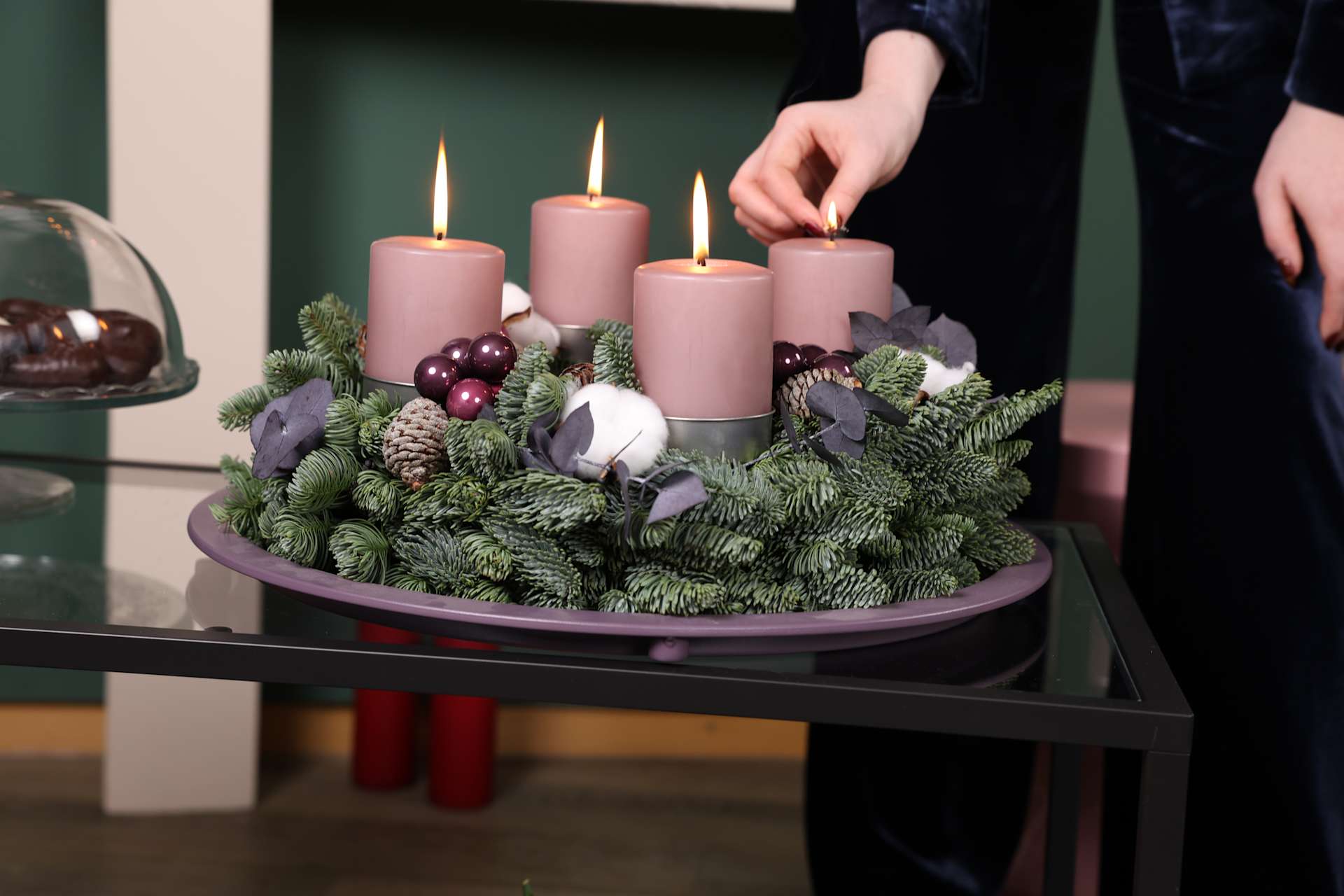 Ein festlicher Adventskranz mit vier rosa Kerzen, umgeben von immergrünen Zweigen, Tannenzapfen und anderen Dekorationselementen, steht auf einem Holzregal vor dunklem Hintergrund.