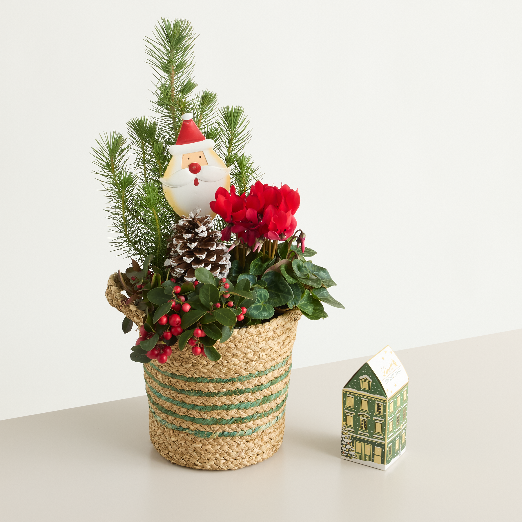 Ein festliches Weihnachtsarrangement mit einer Weihnachtsmannfigur, Tannenzweigen, roten Blumen und einem geflochtenen Korb vor einem schlichten Hintergrund mit einem kleinen Deko-Haus.