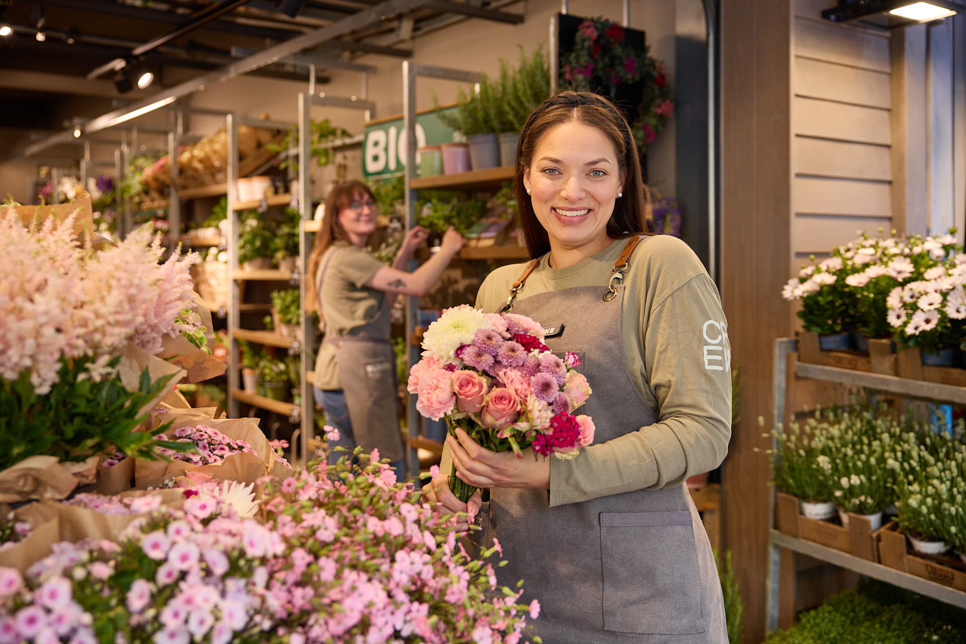 Eine lächelnde Frau in einer geblümten Schürze steht inmitten leuchtender Blumen und Pflanzen in einem Gebäude, das wie ein Blumenladen oder eine Gärtnerei aussieht.
