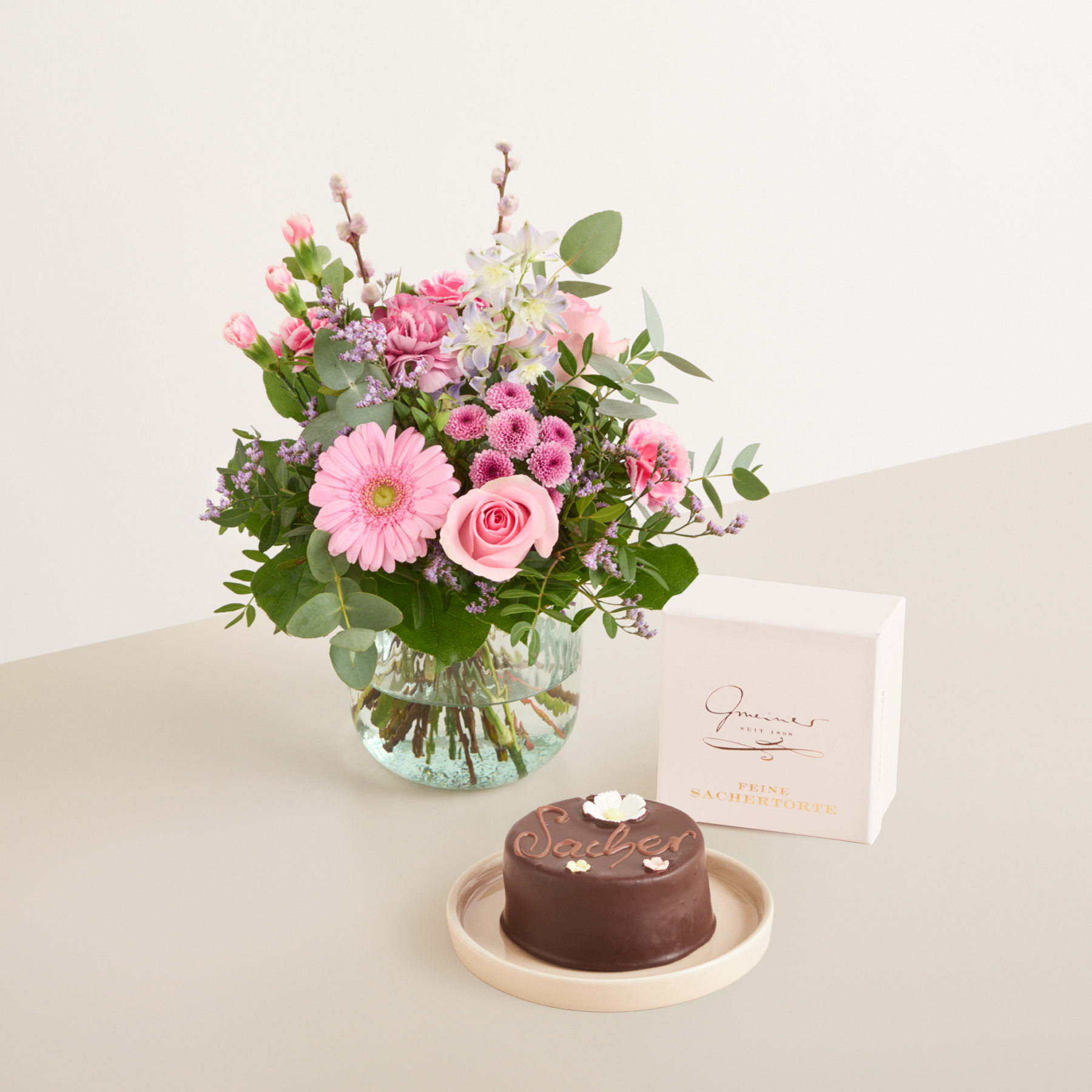 Ein wunderschöner Strauß rosa und weißer Blumen, darunter Rosen und Gerbera, steht in einer Glasvase auf einem Holztisch, dazu ein kleiner Schokoladenkuchen und eine handgeschriebene Notiz.
