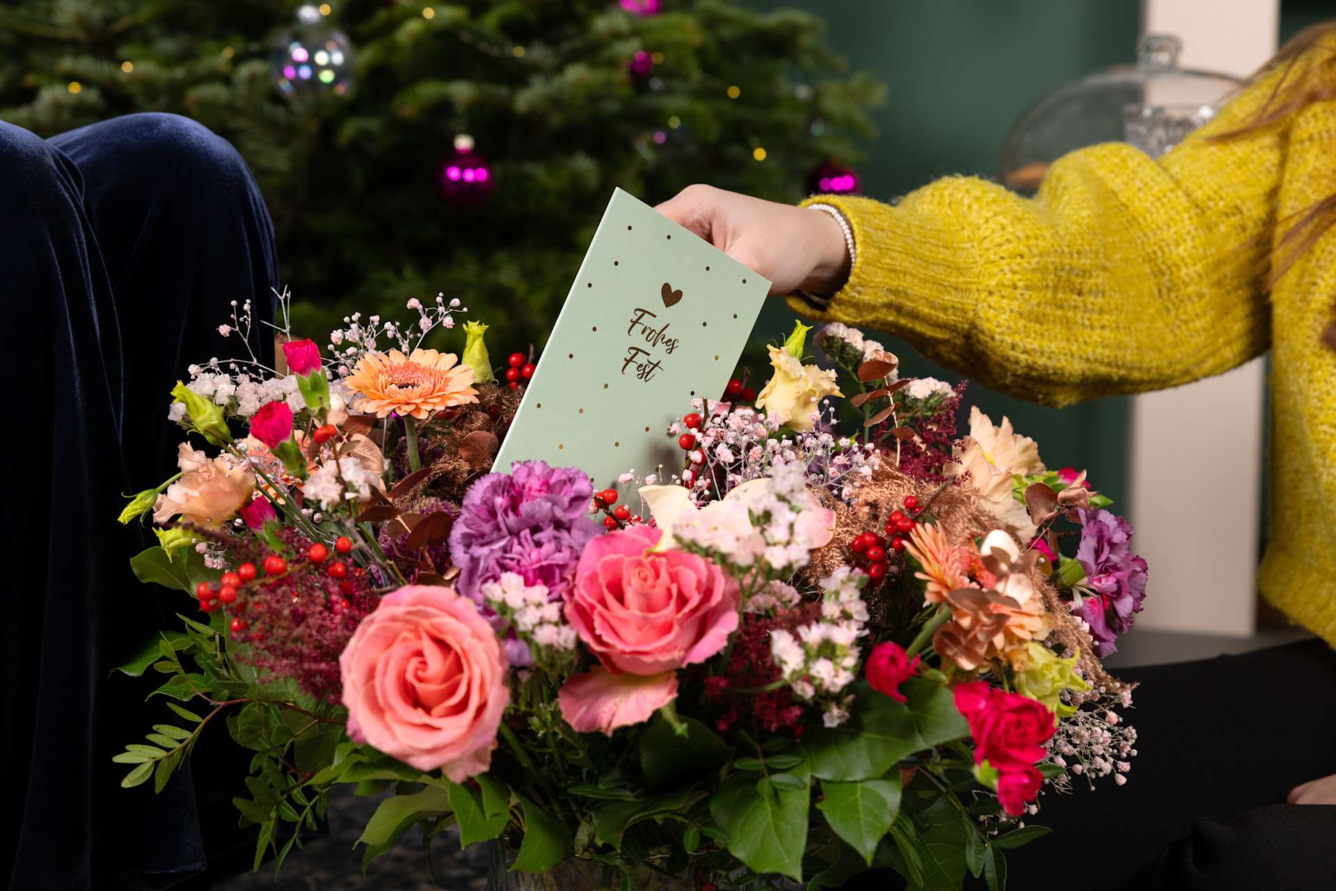 Ein farbenfroher Blumenstrauß mit Rosen, Gänseblümchen und anderen Blüten wird von einer Person in einem gelben Pullover vor dem Hintergrund eines mit funkelnden Lichtern geschmückten Weihnachtsbaums gehalten.
