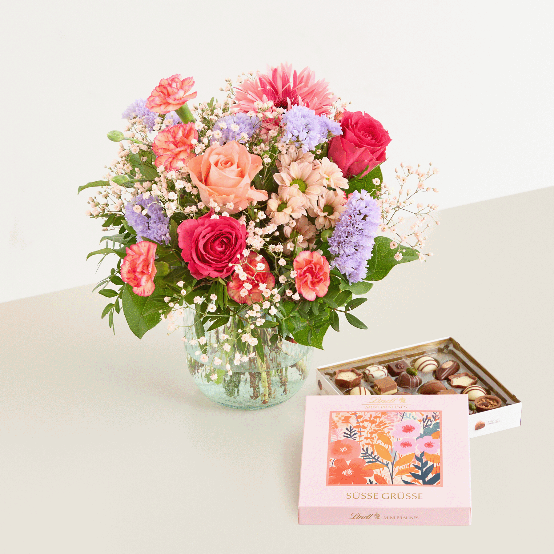 Ein farbenfroher Strauß aus Rosen, Nelken und Gänseblümchen steht in einer Glasvase auf einer hellen Unterlage, daneben eine Schachtel Pralinen.