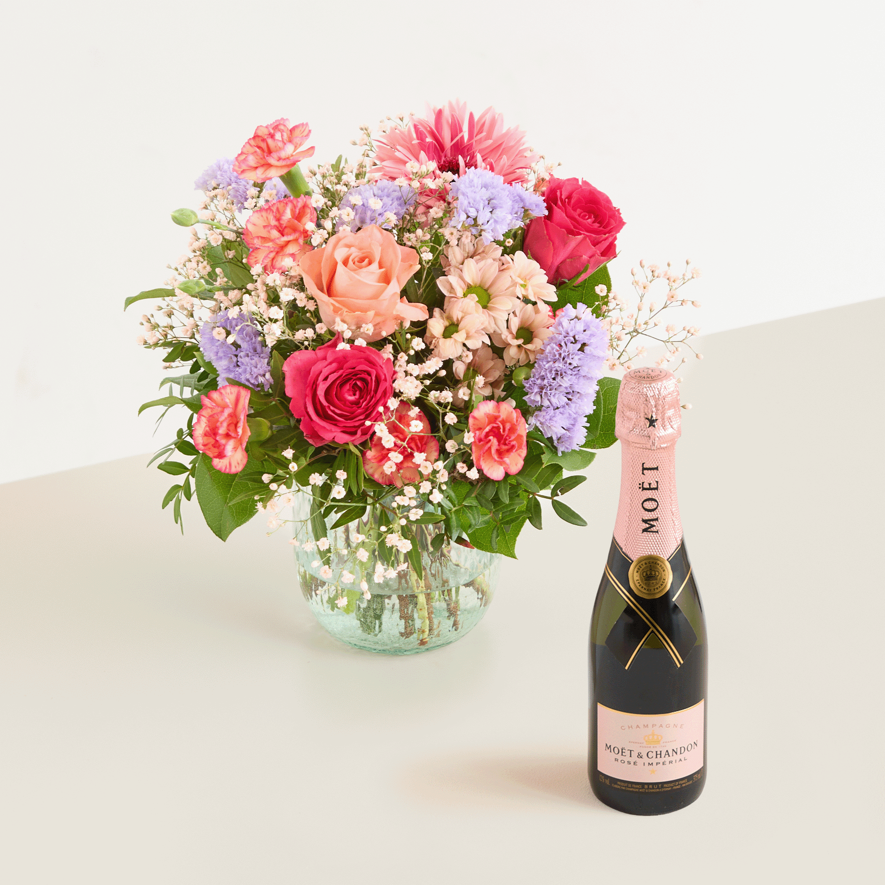 Ein farbenfroher Strauß aus Rosen, Nelken und Gänseblümchen steht in einer Glasvase neben einer Flasche Moët-Champagner auf hellem Hintergrund.