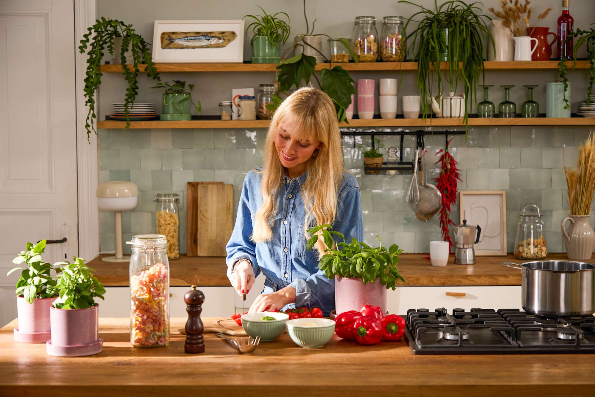 Eine Frau mit blonden Haaren steht in einer Küche, umgeben von verschiedenen Pflanzen, Kochutensilien und anderen Küchengegenständen in Regalen im Hintergrund.