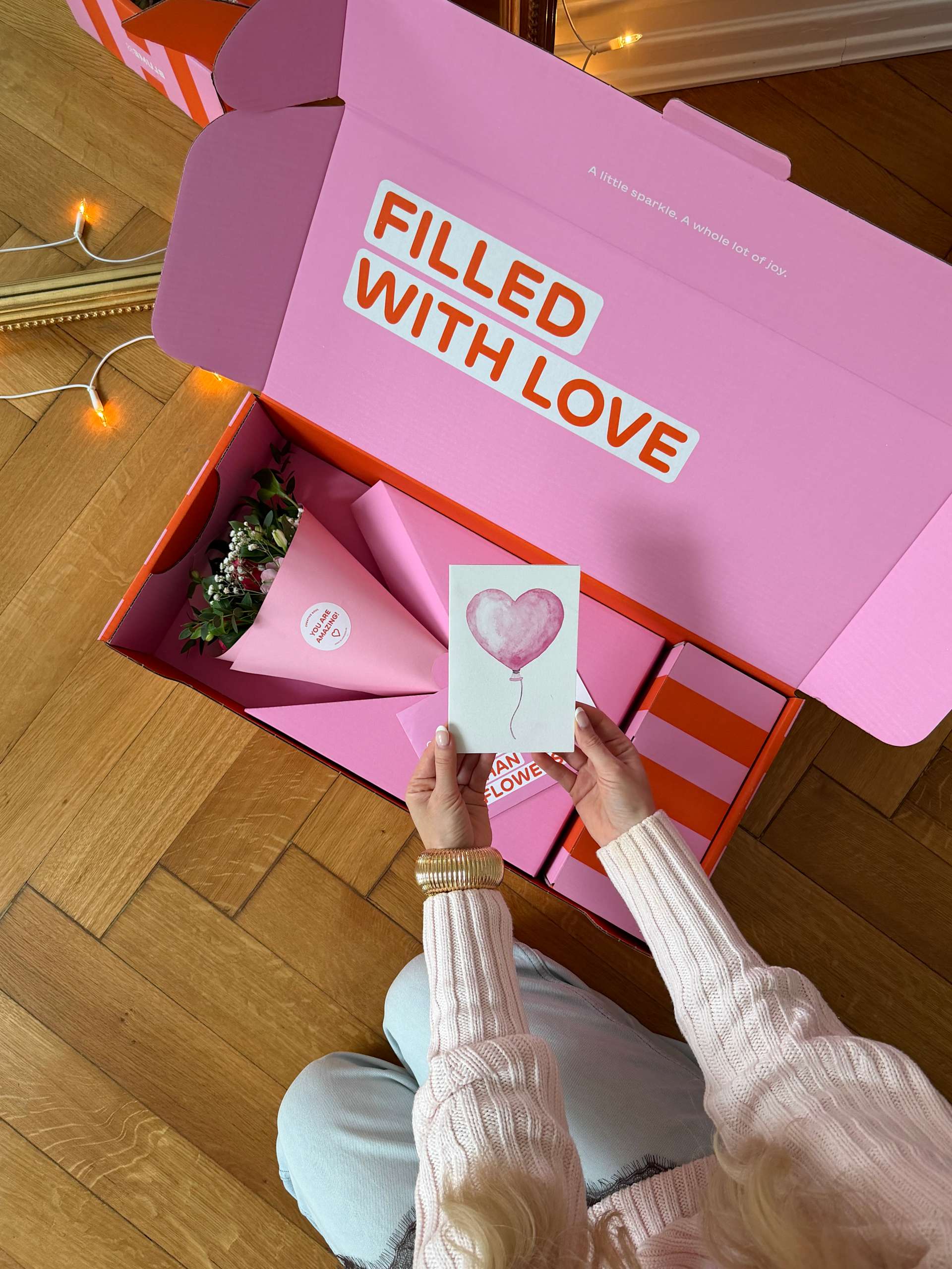 Auf einem Holzboden steht eine große rosa Schachtel mit der Aufschrift „Filled with Love“, in deren Händen eine Karte mit einem Herzsymbol gehalten wird.
