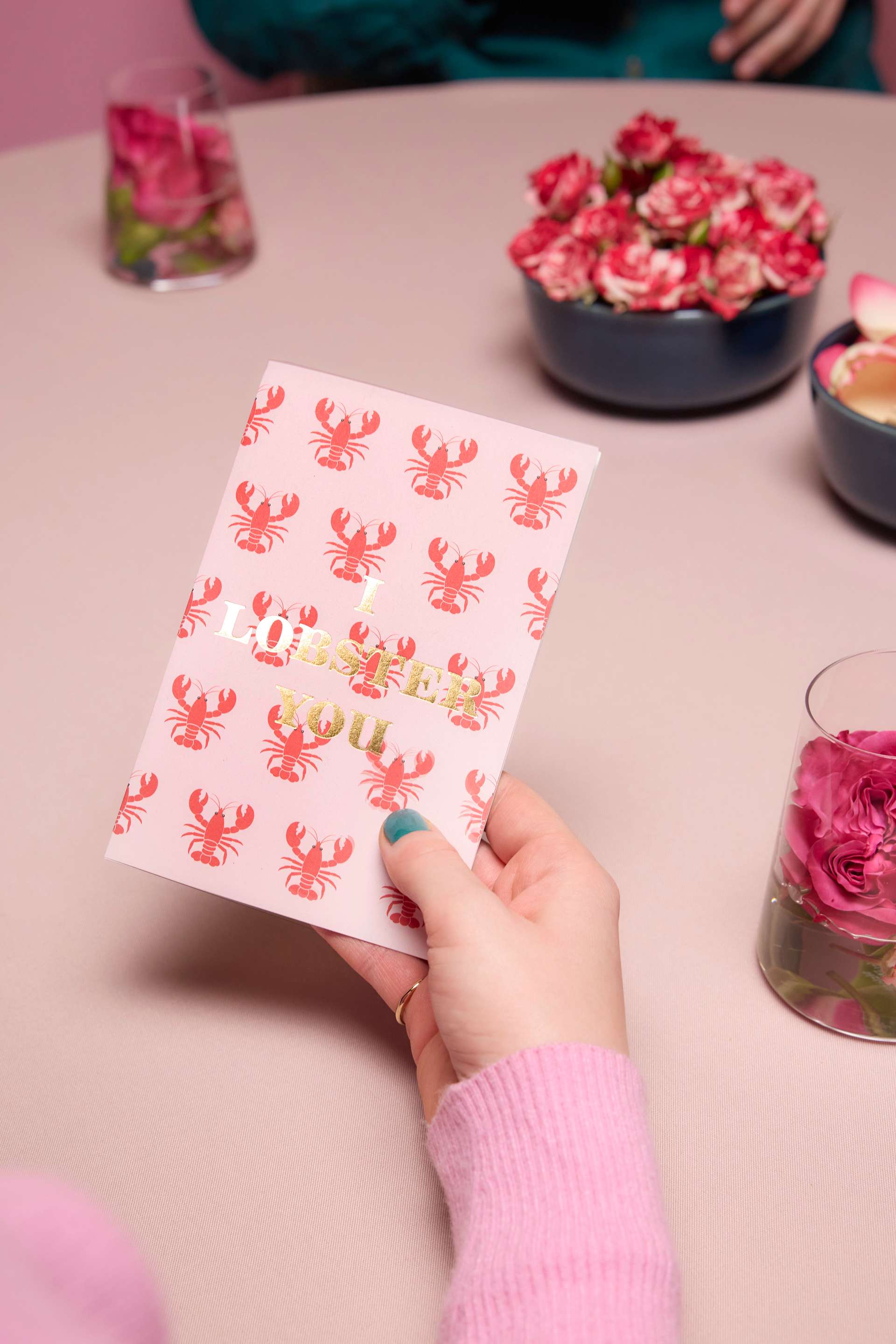 Die Hände einer Person halten ein rot-weiß gemustertes Notizbuch auf einem rosa Tisch; im Hintergrund sind eine Vase mit rosa Blumen und andere Dekorationsgegenstände zu sehen.