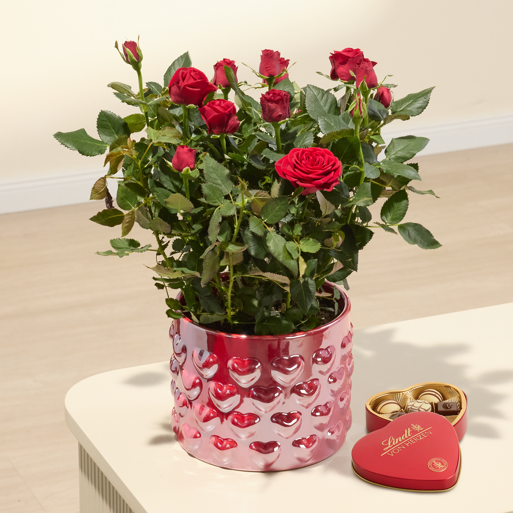Auf einer Holzoberfläche steht ein leuchtend roter Rosenstrauß in einem dekorativen rosa Topf, daneben eine herzförmige Pralinenschachtel.