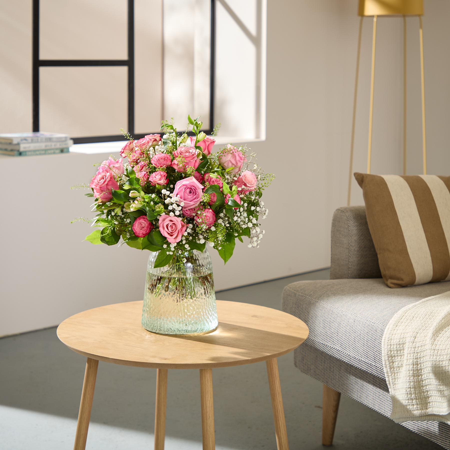 Eine junge Frau mit langen braunen Haaren hält vor einem schlichten weißen Hintergrund einen leuchtenden Strauß aus rosa Rosen und anderen Blumen.