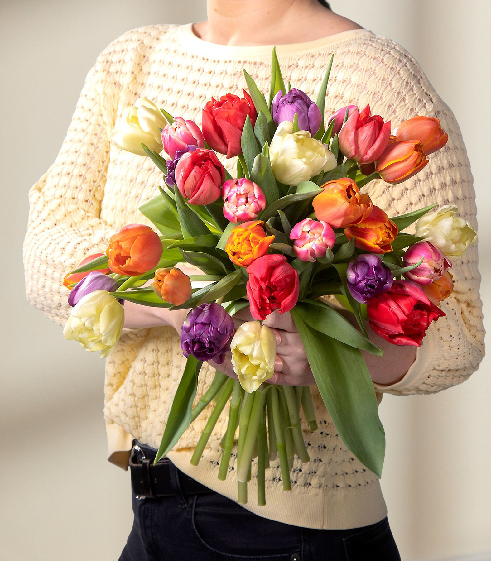 Im Vordergrund wird ein farbenfroher Strauß aus Tulpen und anderen Frühlingsblumen gehalten, im Hintergrund ist eine Person in einem cremefarbenen Pullover zu sehen.