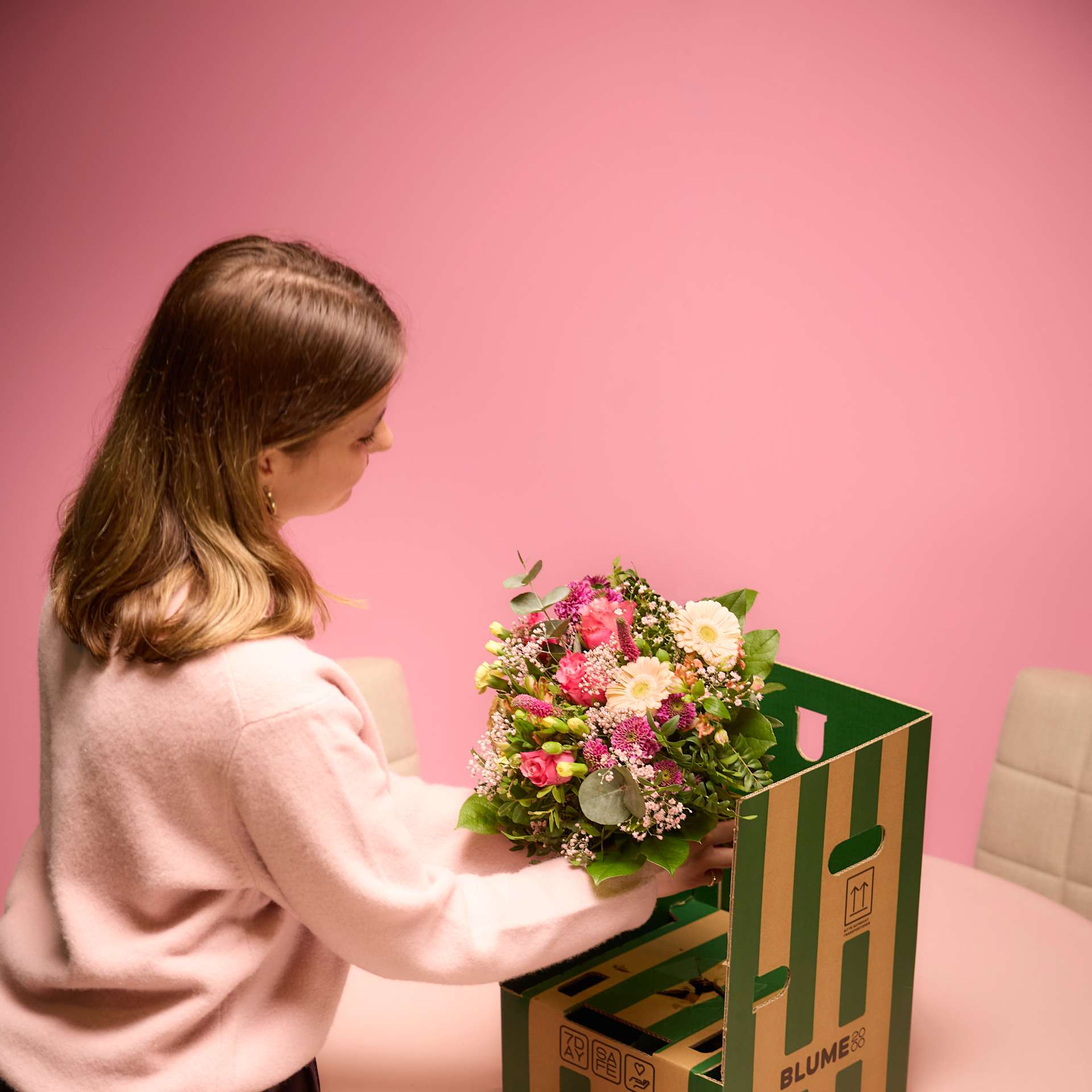 Eine junge Frau in einem rosa Pullover hält einen leuchtenden Blumenstrauß vor einem einfarbig rosa Hintergrund.