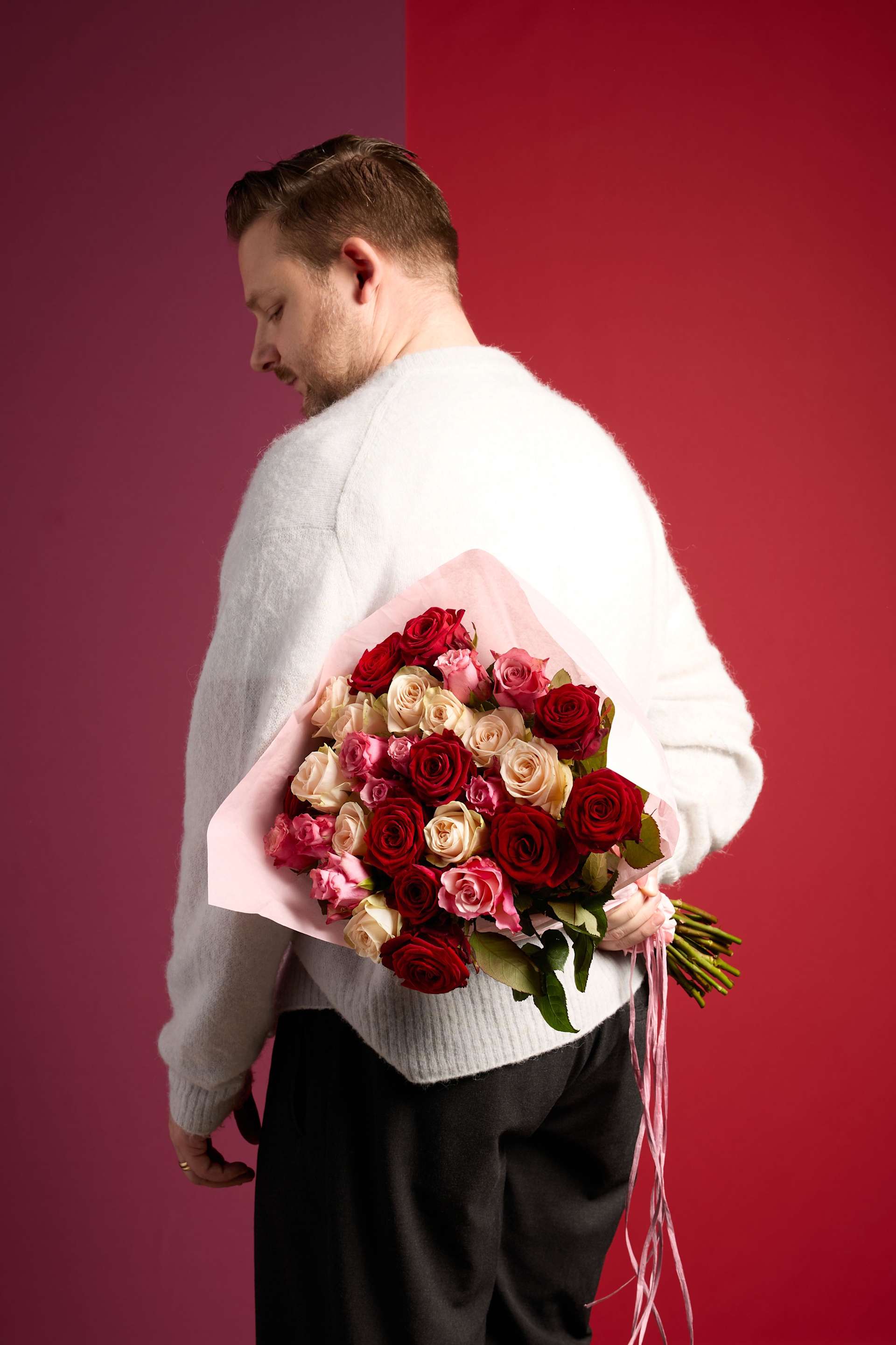 Ein Mann in einem weißen Pullover hält einen Strauß roter und weißer Rosen vor einem leuchtend roten Hintergrund.