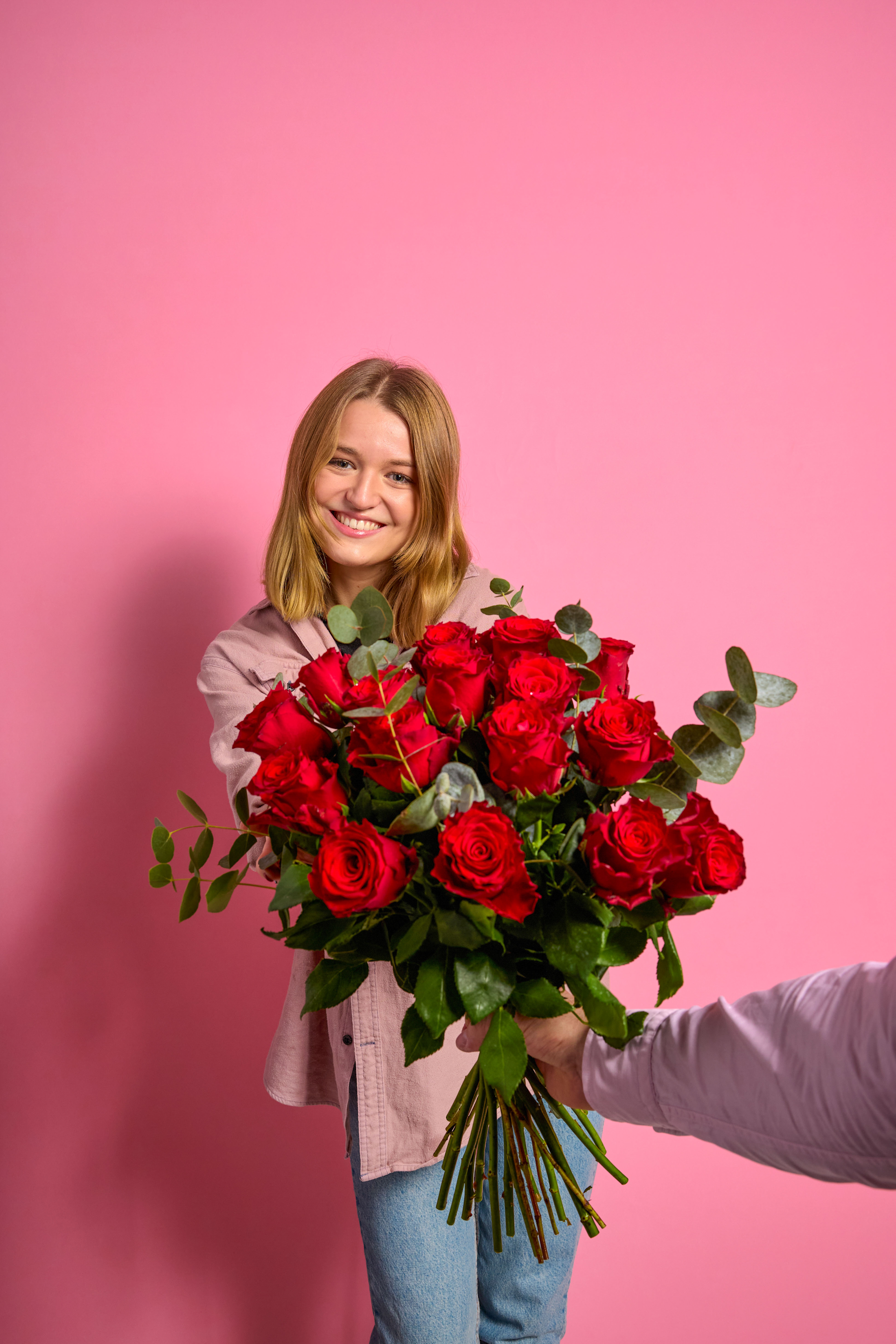 Eine lächelnde junge Frau hält einen großen Strauß leuchtend roter Rosen vor einem hellrosa Hintergrund.