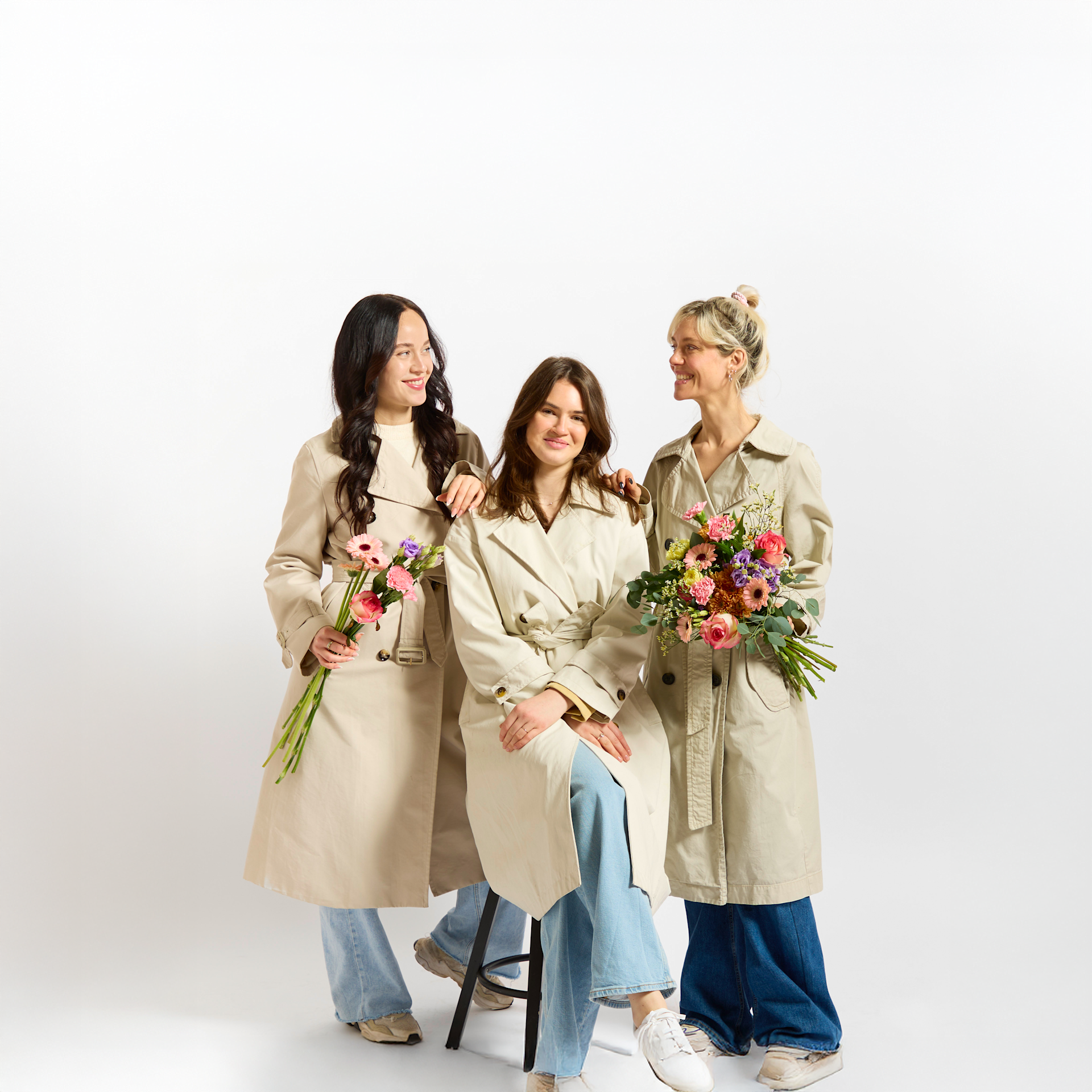 Drei Frauen in Freizeitkleidung stehen zusammen und halten Blumensträuße vor einem schlichten weißen Hintergrund.