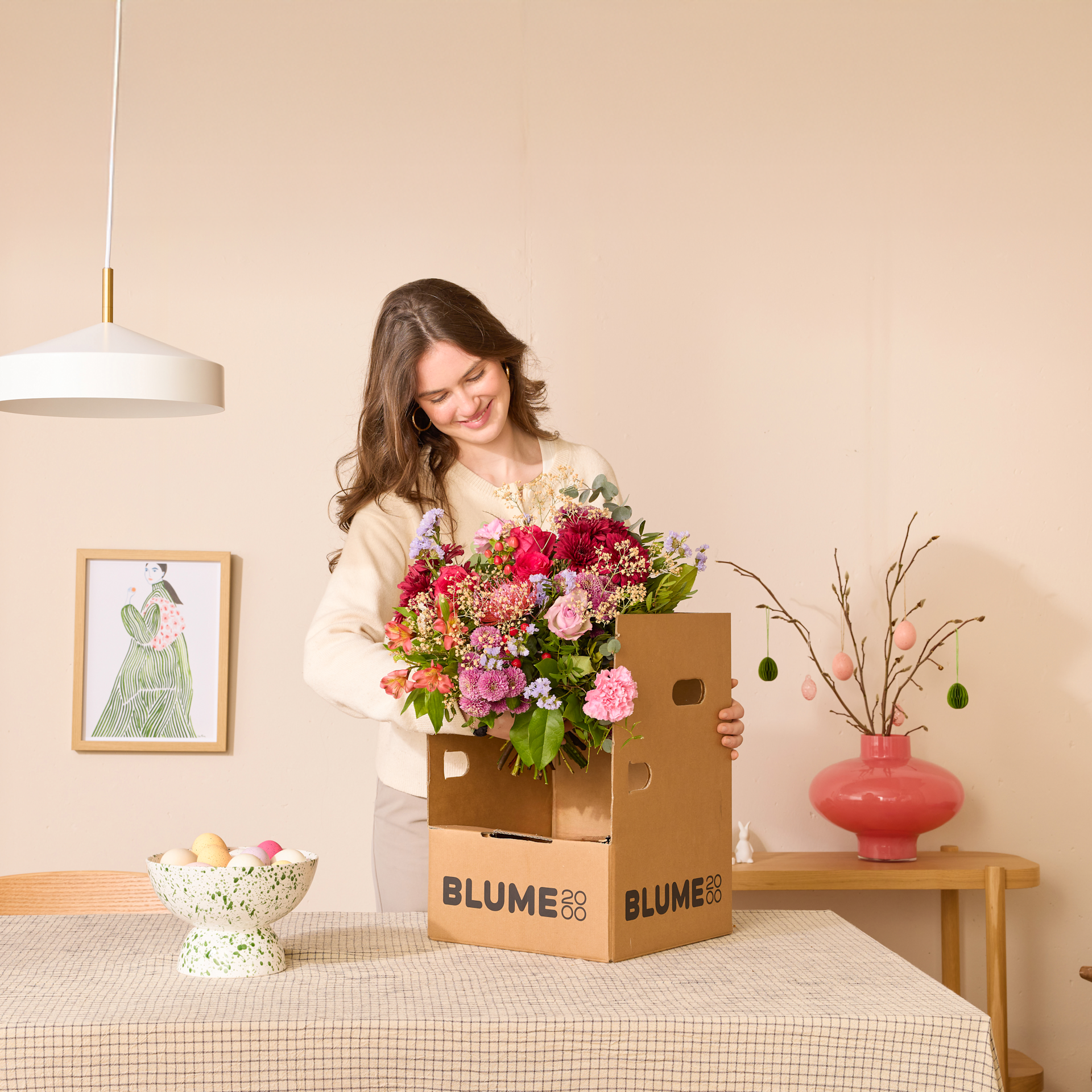 Eine junge Frau mit langen braunen Haaren hält einen großen Strauß bunter Blumen in der Hand und steht in einem gemütlichen Zimmer. Auf einem Tisch stehen ein gerahmtes Kunstwerk, eine Vase und Pappkartons mit der Aufschrift „Blume“.