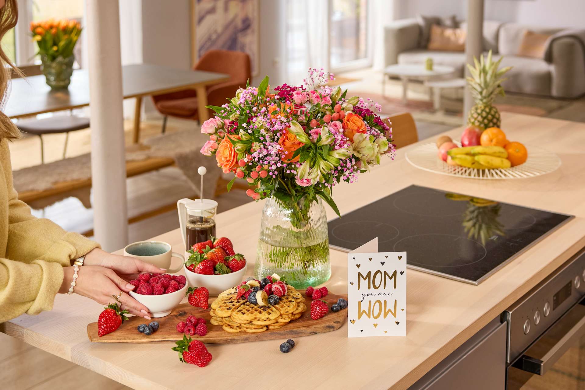 Ein bunter Blumenstrauß, eine Platte mit frischem Obst und ein handgeschriebenes Schild mit der Aufschrift „Mama“ schmücken eine gemütliche Wohnzimmeratmosphäre mit bequemen Möbeln und natürlichem Licht.