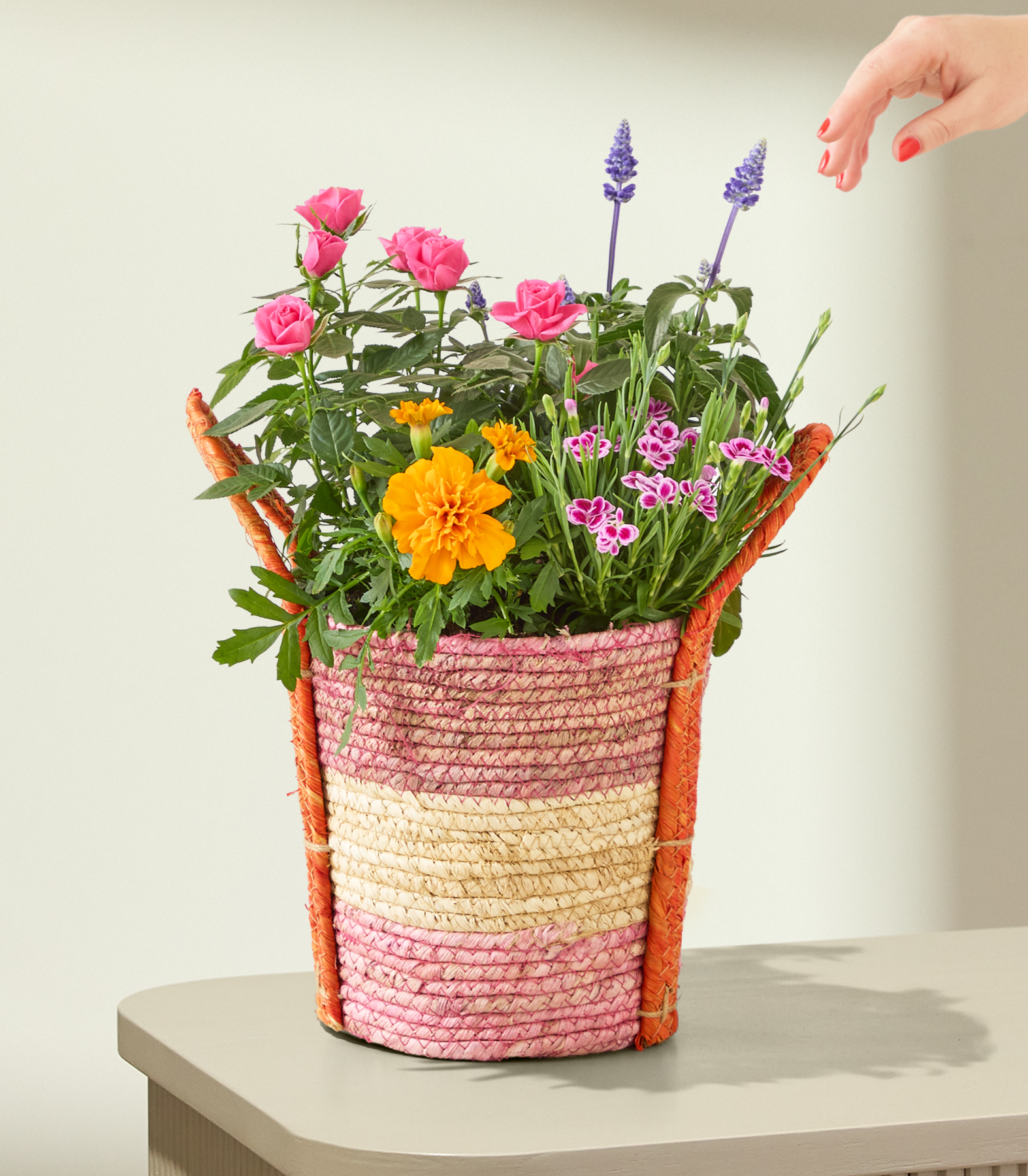 Auf einem Tisch steht ein bunter, geflochtener Korb, der überquillt von rosa Rosen, Ringelblumen und lila Salbei; eine Hand streckt sich vor einem schlichten Hintergrund nach den Blumen aus.