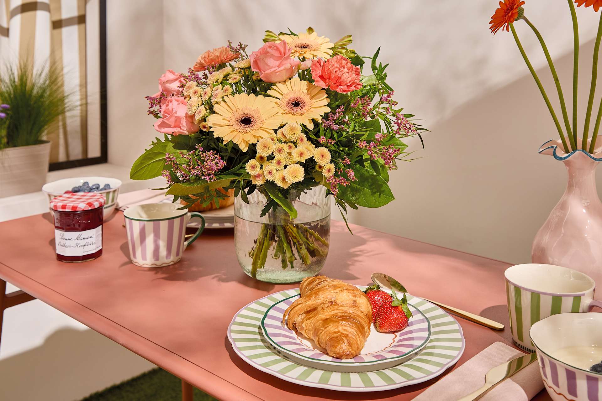 Auf einem rosa Tisch steht ein bunter Blumenstrauß, im Vordergrund sind ein Croissant und Erdbeeren zu sehen, im Hintergrund eine Vase mit orangefarbenen Blumen und eine Topfpflanze.