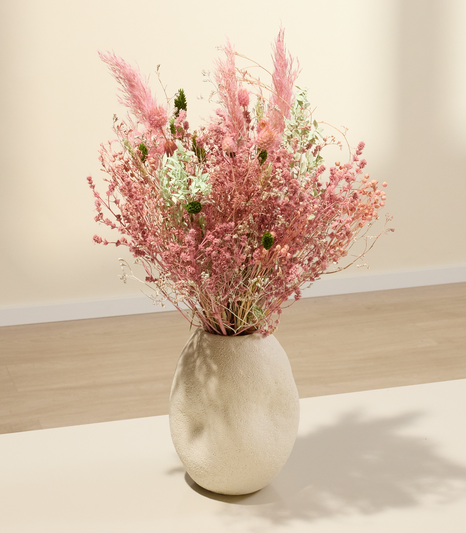 Eine strukturierte, cremefarbene Vase beherbergt einen Strauß zarter rosa und grüner Trockenblumen vor einem sanft beleuchteten, neutralen Hintergrund.