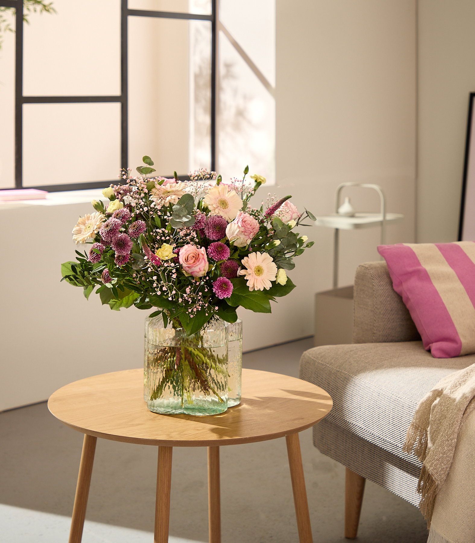 Im Vordergrund steht auf einem Holztisch ein leuchtender Strauß rosa und violetter Blumen in einer Glasvase, im sanft beleuchteten Hintergrund sind ein Sofa und ein Fenster zu sehen.