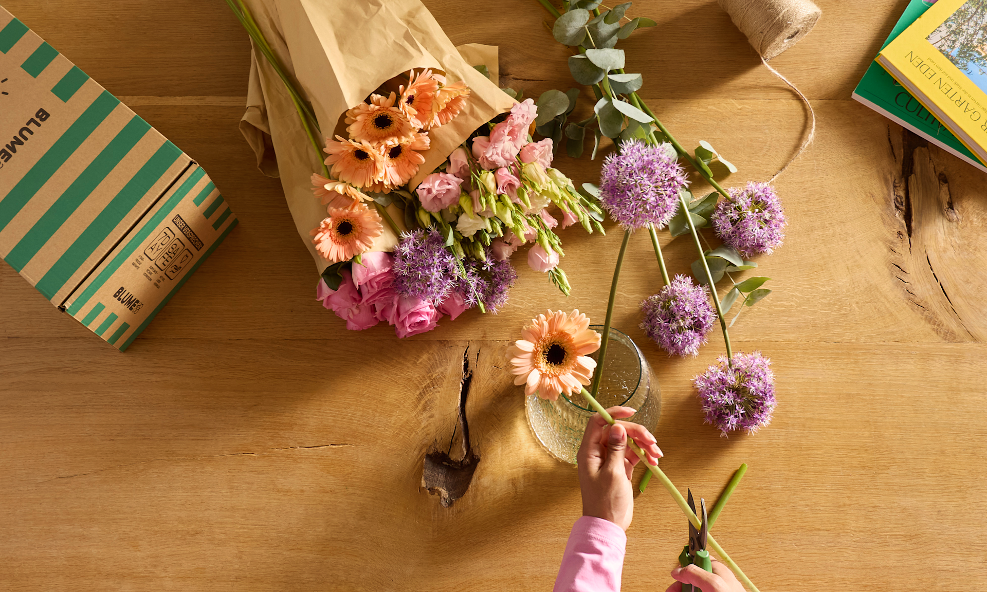 Im Vordergrund stutzen die Hände einer Person eine rosa Gerbera, während im Hintergrund auf einem Holztisch ein Strauß bunter Blumen, eingewickelt in braunes Papier, und lila Zierlauch in einer Vase stehen.