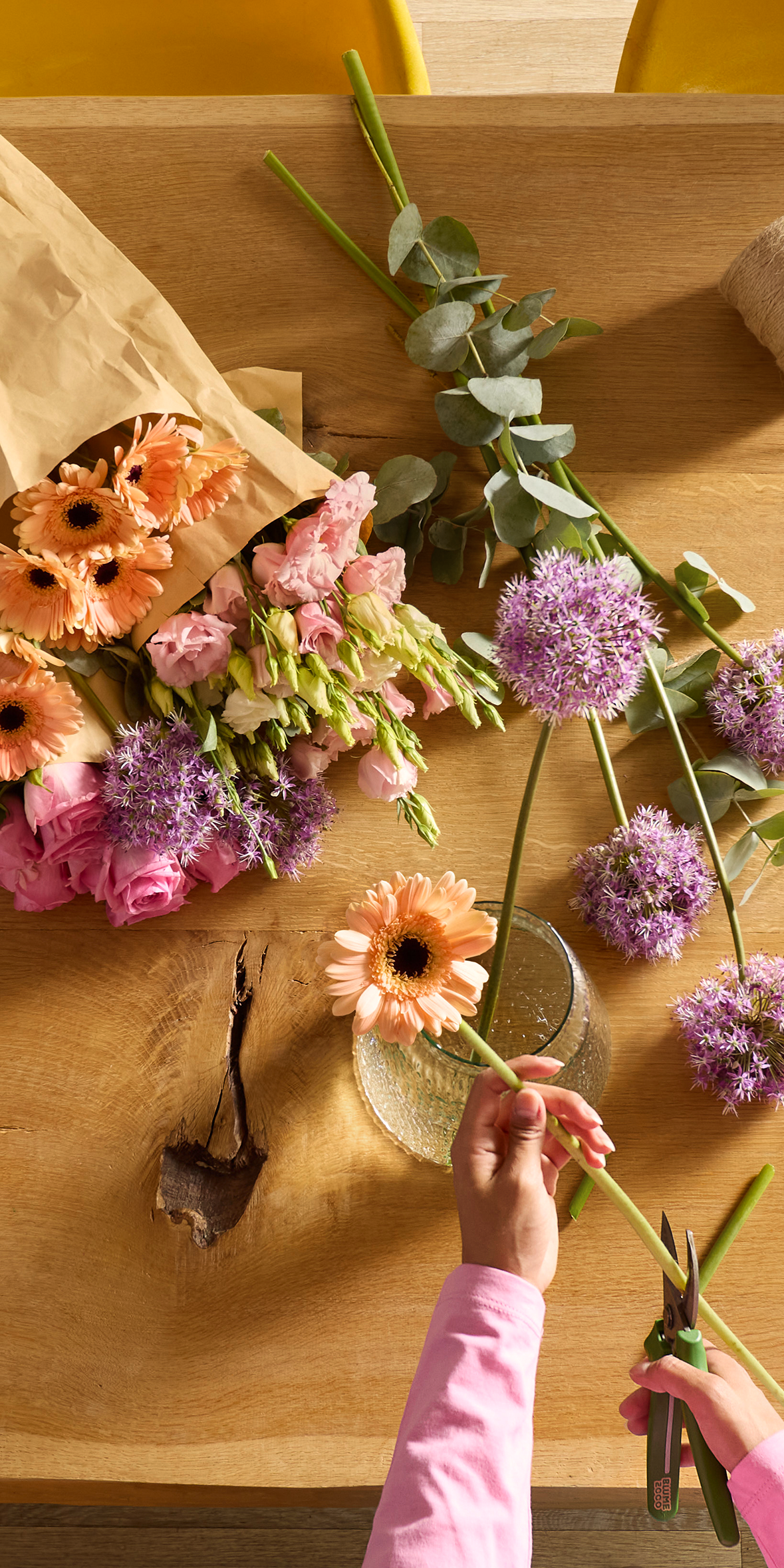 Im Vordergrund stutzen die Hände einer Person eine rosa Gerbera, während im Hintergrund auf einem Holztisch ein Strauß bunter Blumen, eingewickelt in braunes Papier, und lila Zierlauch in einer Vase stehen.