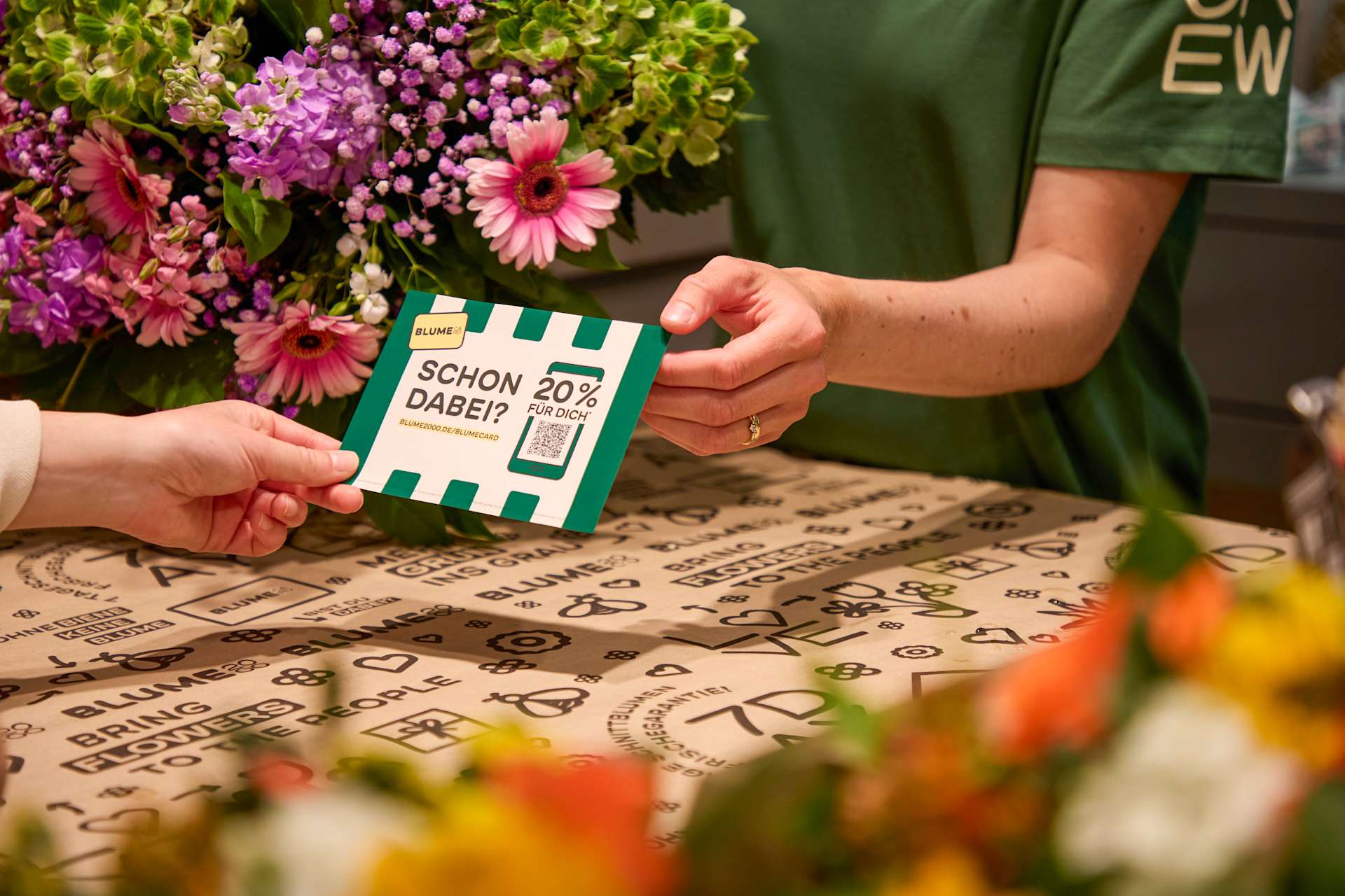 Eine Hand reicht einer anderen Hand vor einem Blumenstrauß eine Rabattkarte mit QR-Code; im verschwommenen Hintergrund ist eine Person in einem grünen Hemd zu erkennen.