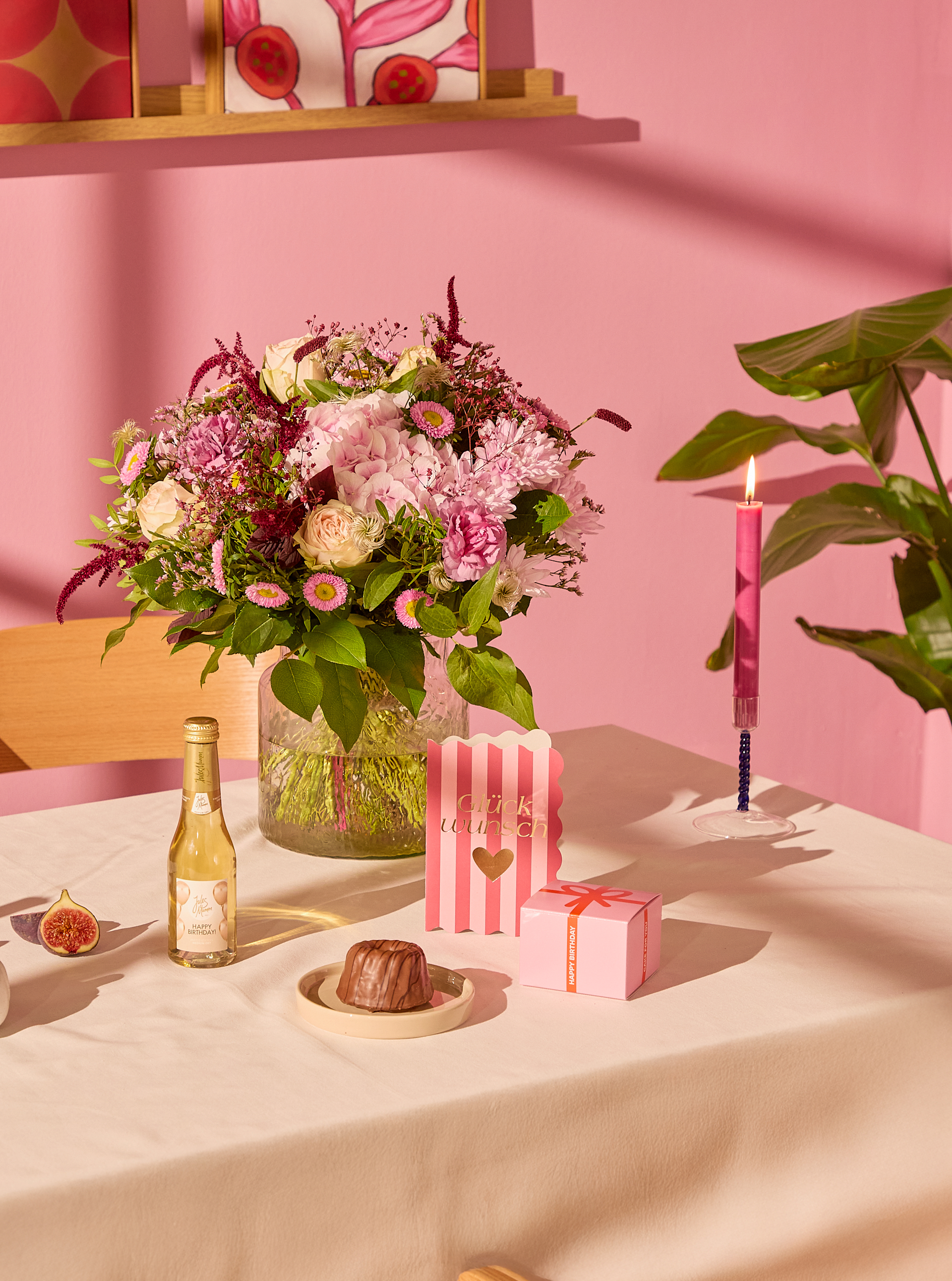 Auf einem Tisch steht ein Strauß rosa Blumen zusammen mit einem kleinen Kuchen und Geschenken, während im Hintergrund vor einer rosa Wand eine brennende rosa Kerze und eine Pflanze zu sehen sind. (Beschreibungstext KI-generiert)