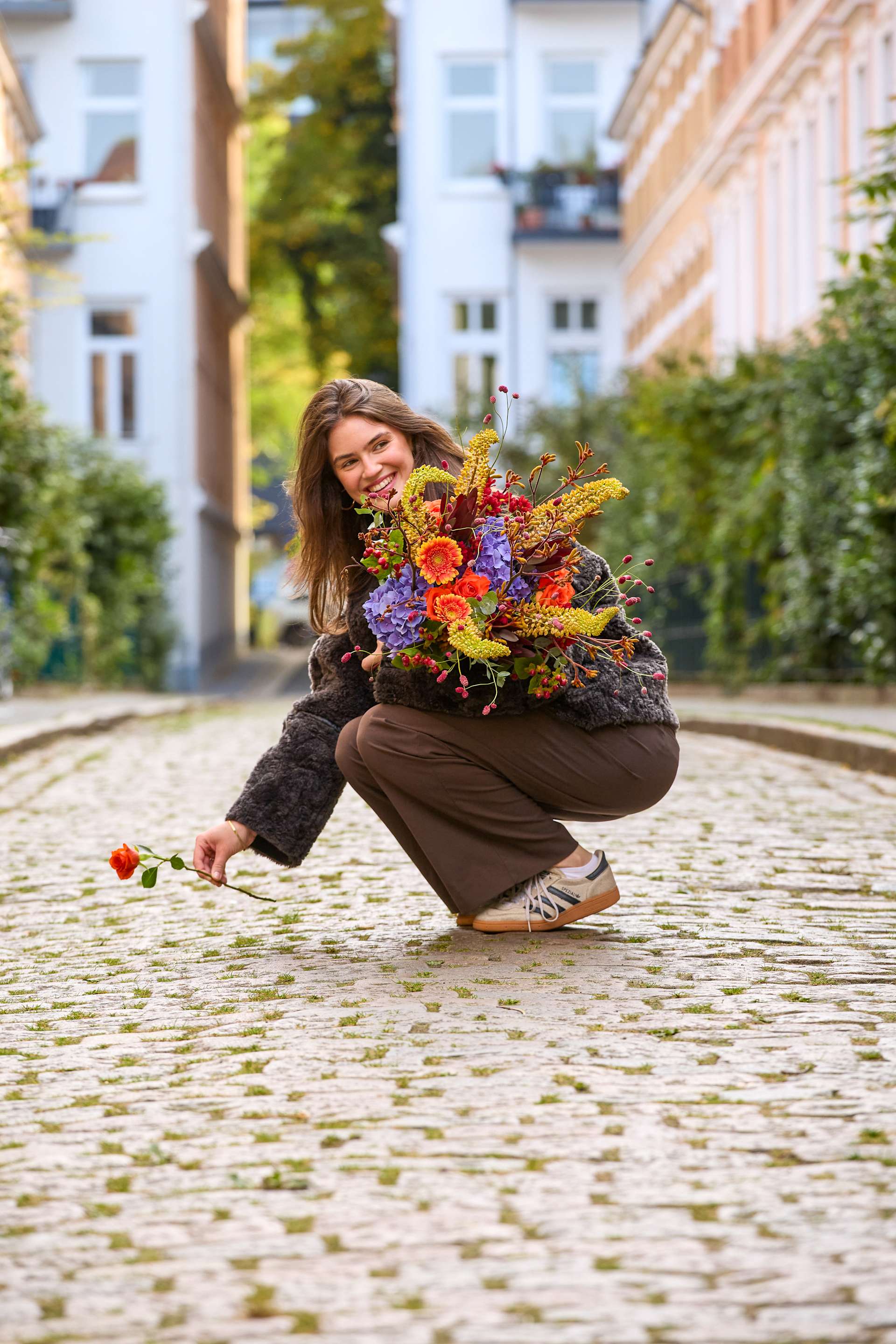 Eine lächelnde Frau in brauner Kleidung hockt auf einer Kopfsteinpflasterstraße und hält einen bunten Blumenstrauß in der Hand; im Hintergrund sind Gebäude zu sehen. (Beschreibungstext KI-generiert)