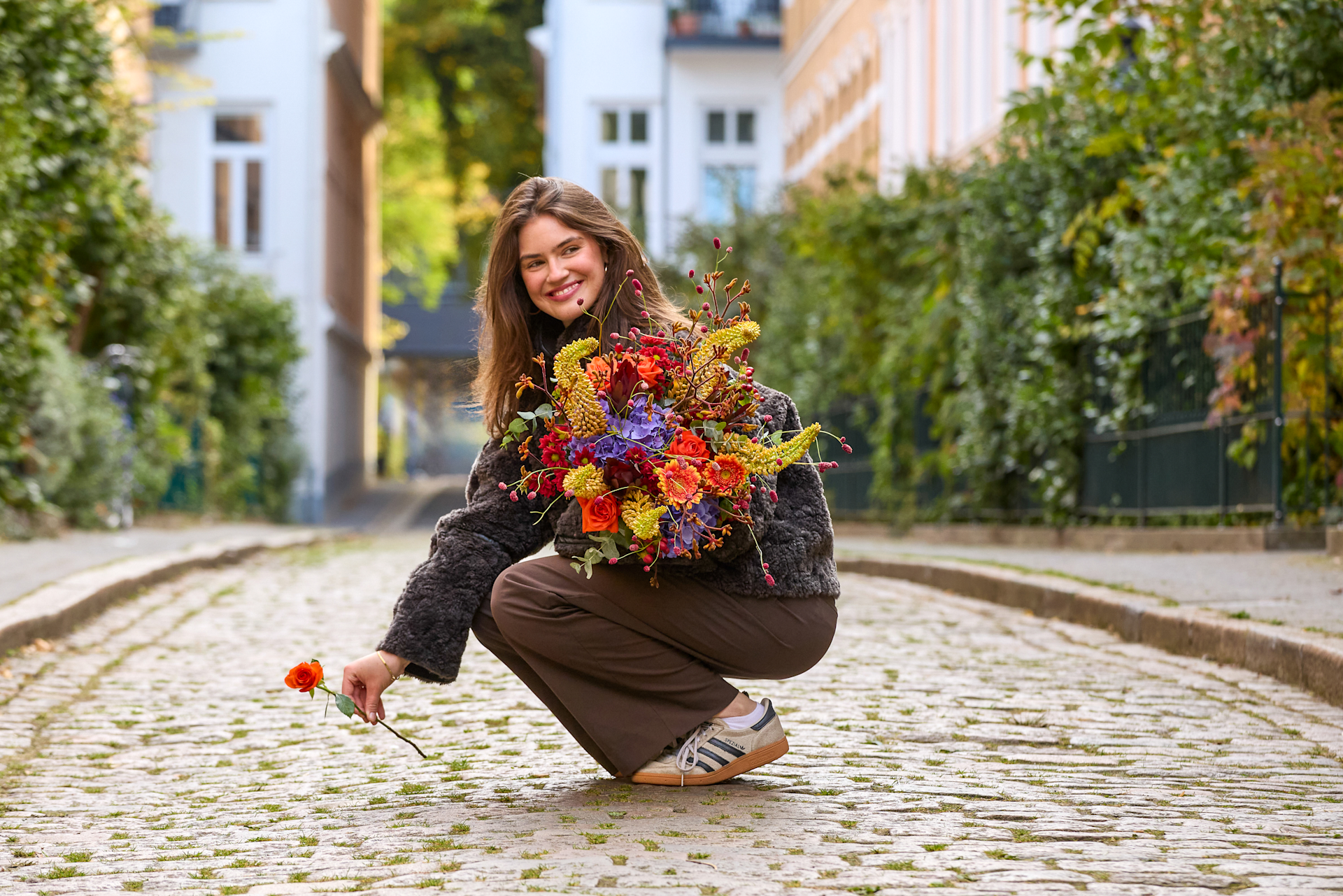 Eine lächelnde Frau hockt auf einem Kopfsteinpflaster, hält einen farbenprächtigen Blumenstrauß in der Hand; im Hintergrund erheben sich Gebäude und Grünanlagen. (Beschreibungstext KI-generiert)