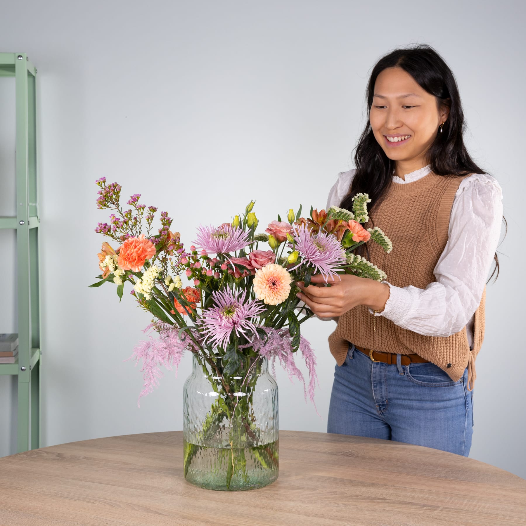 Eine lächelnde junge Frau in legerer Kleidung arrangiert einen farbenfrohen Strauß bunter Blumen in einer Glasvase auf einem Holztisch, im Hintergrund ist ein grüner Schrank zu sehen.